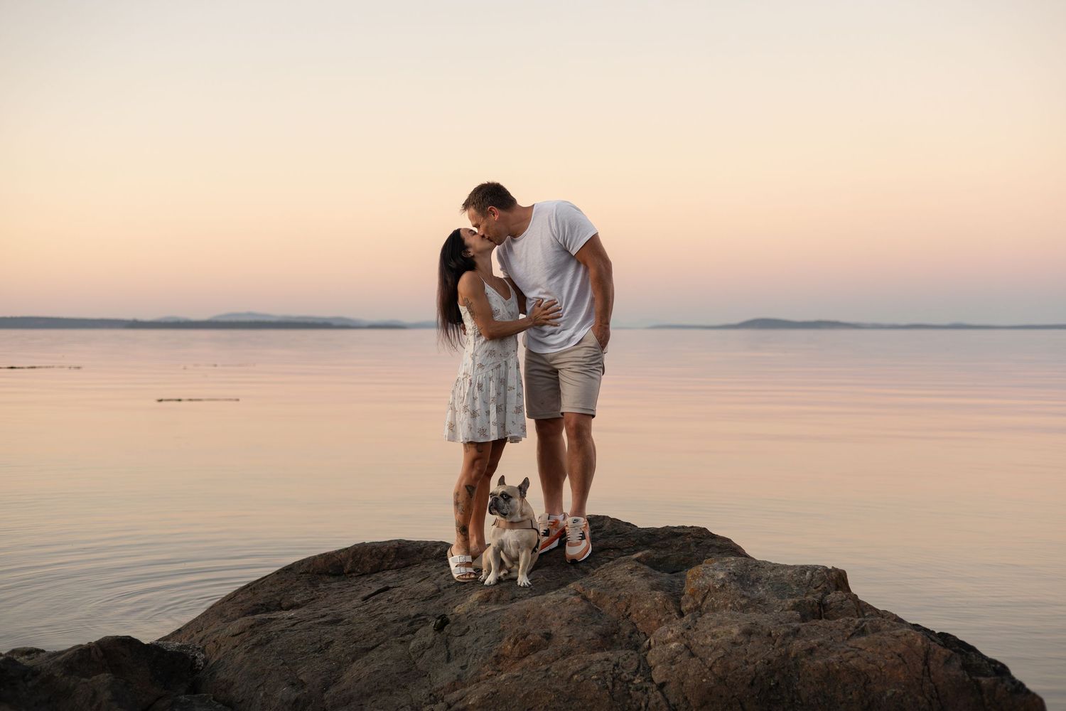 A couple standing on a rock by the ocean kissing each other at sunset. Their dog sits in front of them.