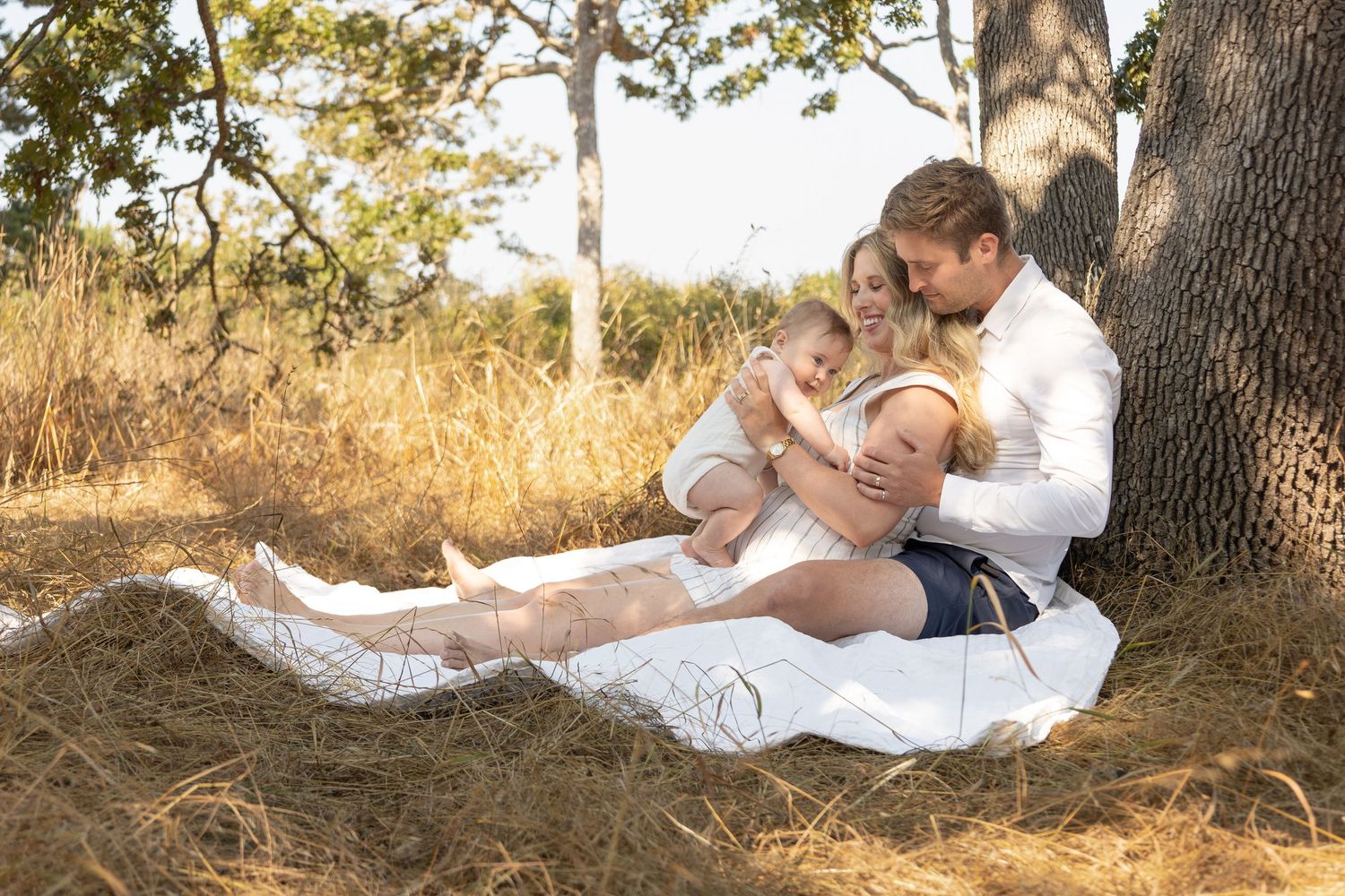 Two parents snuggle on a blanket in a field under a tree with their new baby.