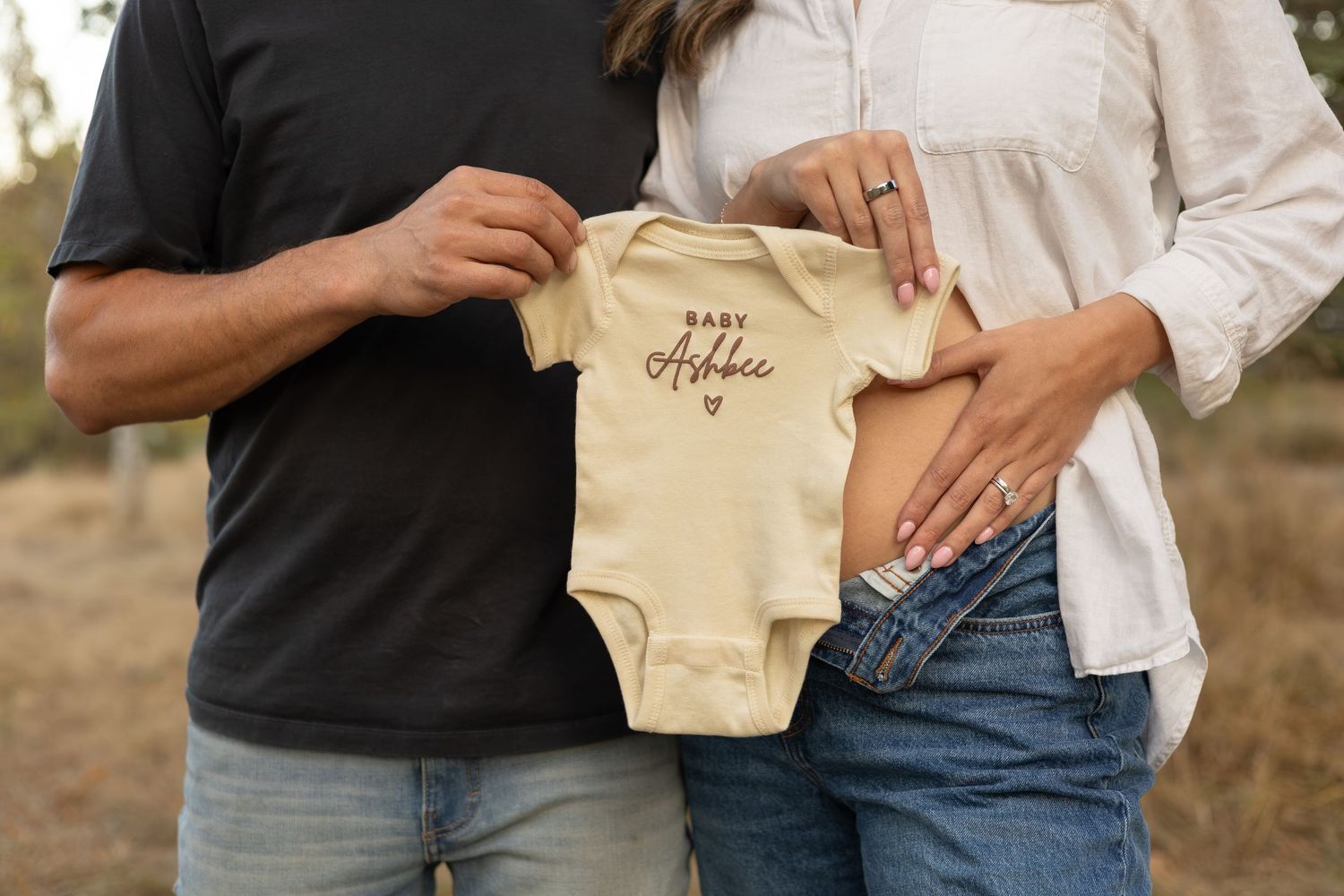 A couple holds up a baby onesie while the mom holds her belly to announce her pregnancy.