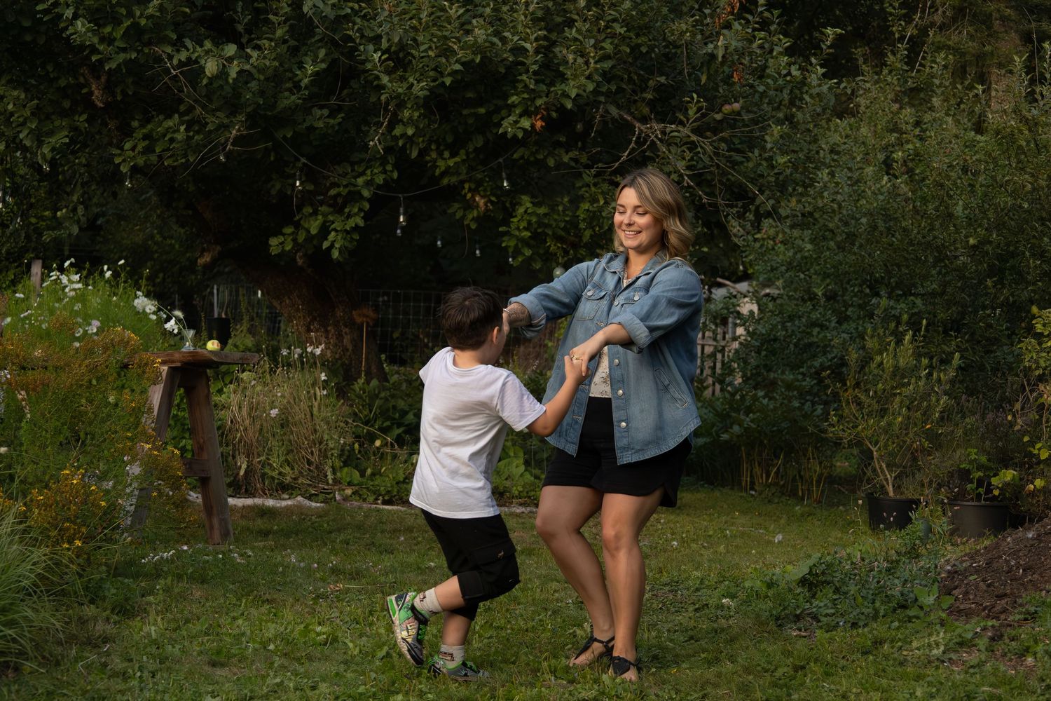 A mom smiles at her young son while they hold hands and dance together in a garden.
