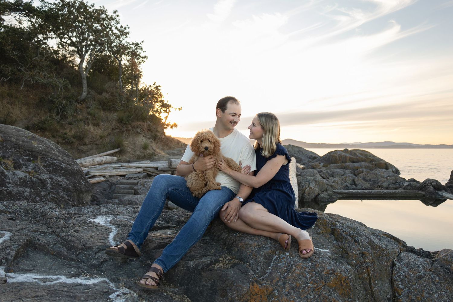 A young couple sits on a large rock beside the ocean at sunset. They smile and look at each other while holding a new puppy in their laps.
