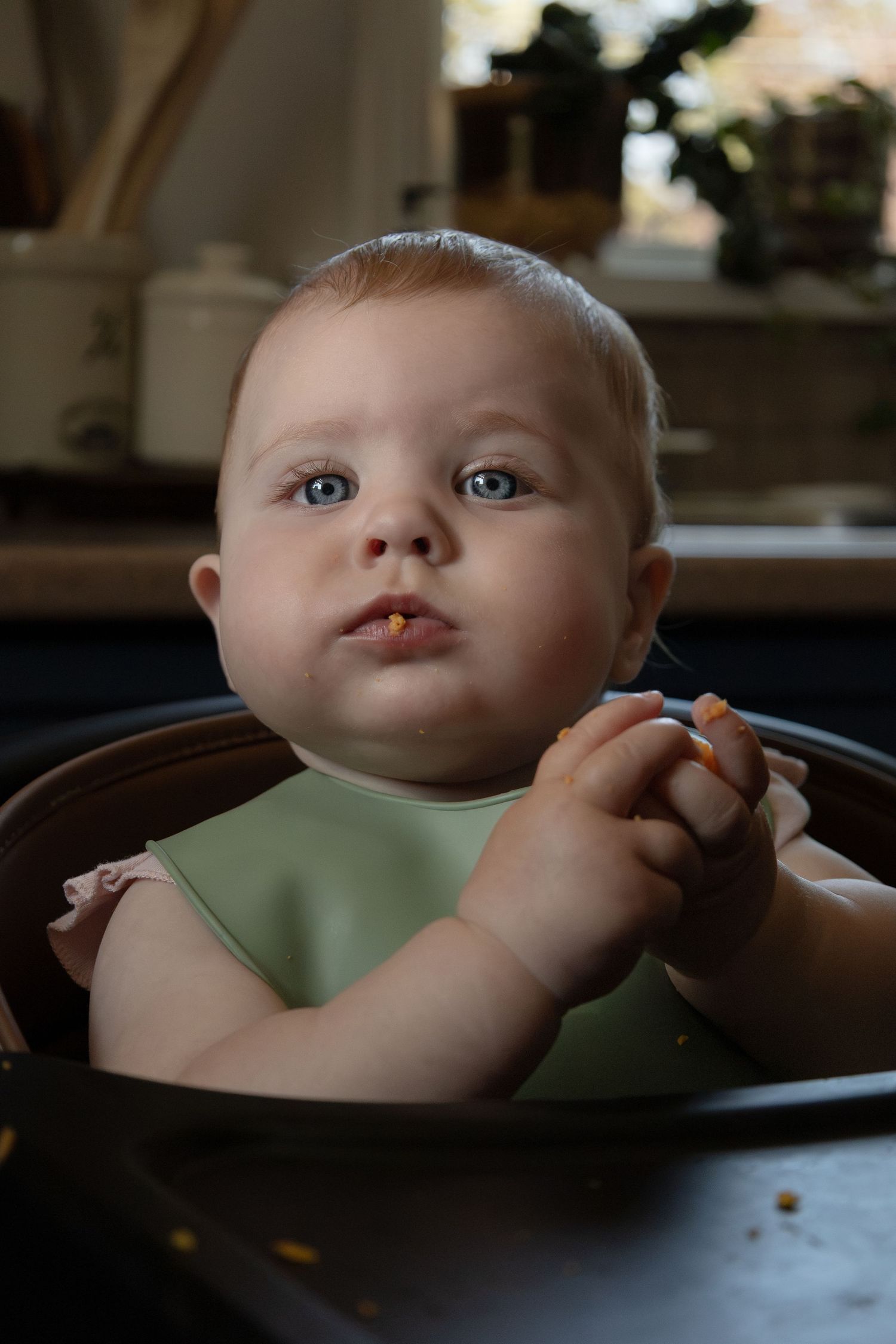 A baby sits in a high chair eating food while looking at the camera.