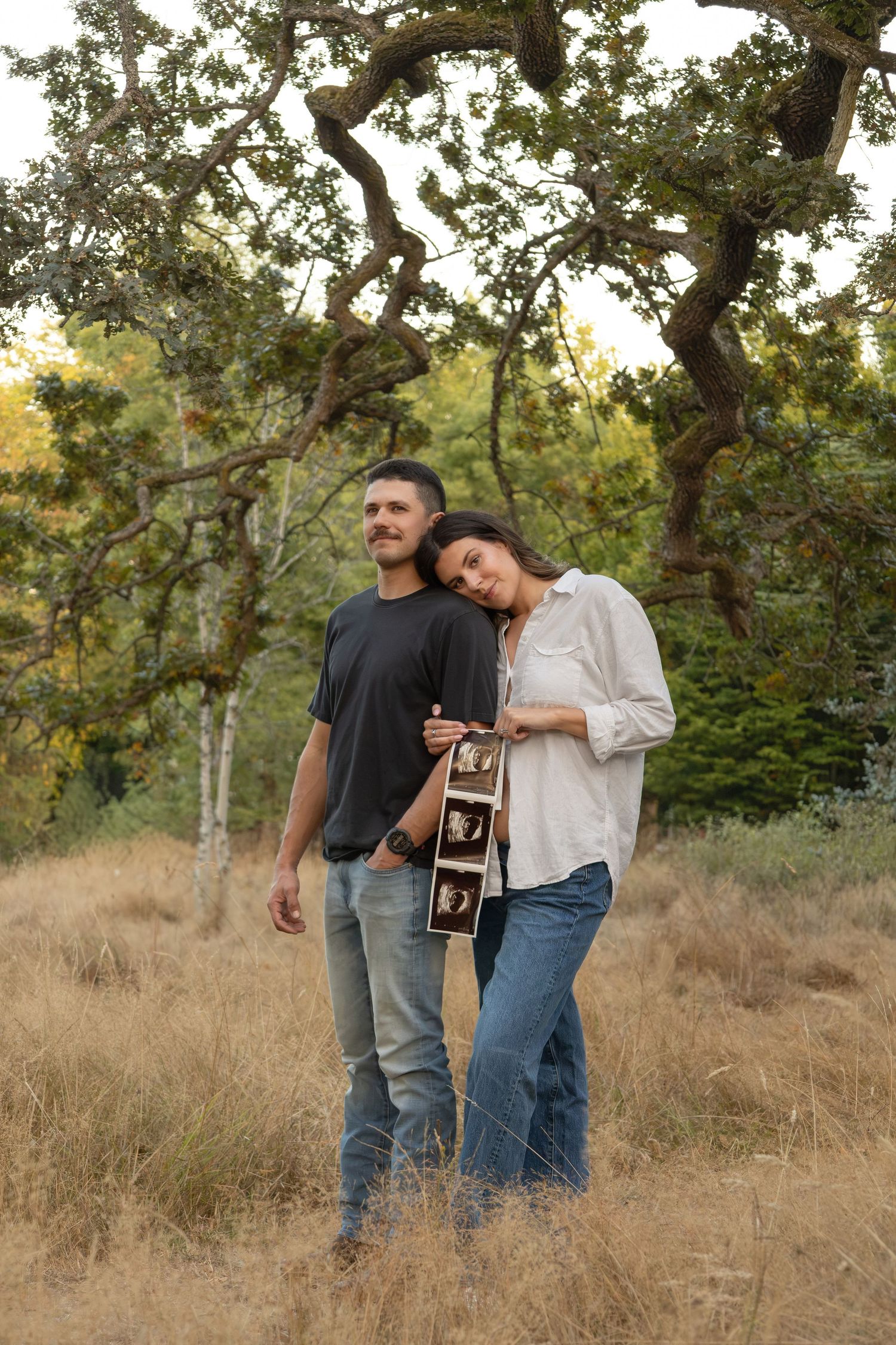 A couple stands together in a field looking off into the distance. The women leans her head on the mans shoulder and she holds a sonogram.