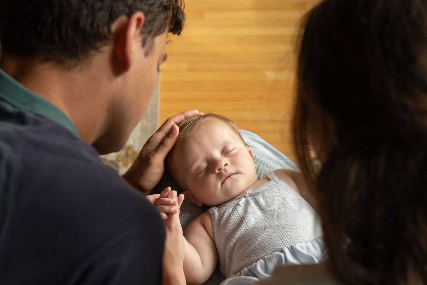 Two parents sit beside each other looking down at their new baby sleeping in their lap. The mom holds the baby's hand.