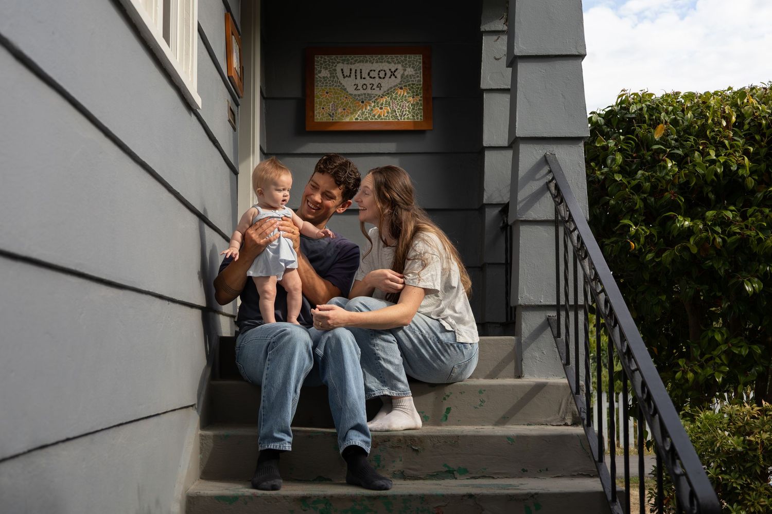 Young parents sit in the sun on the front steps outside their home playing with their baby.