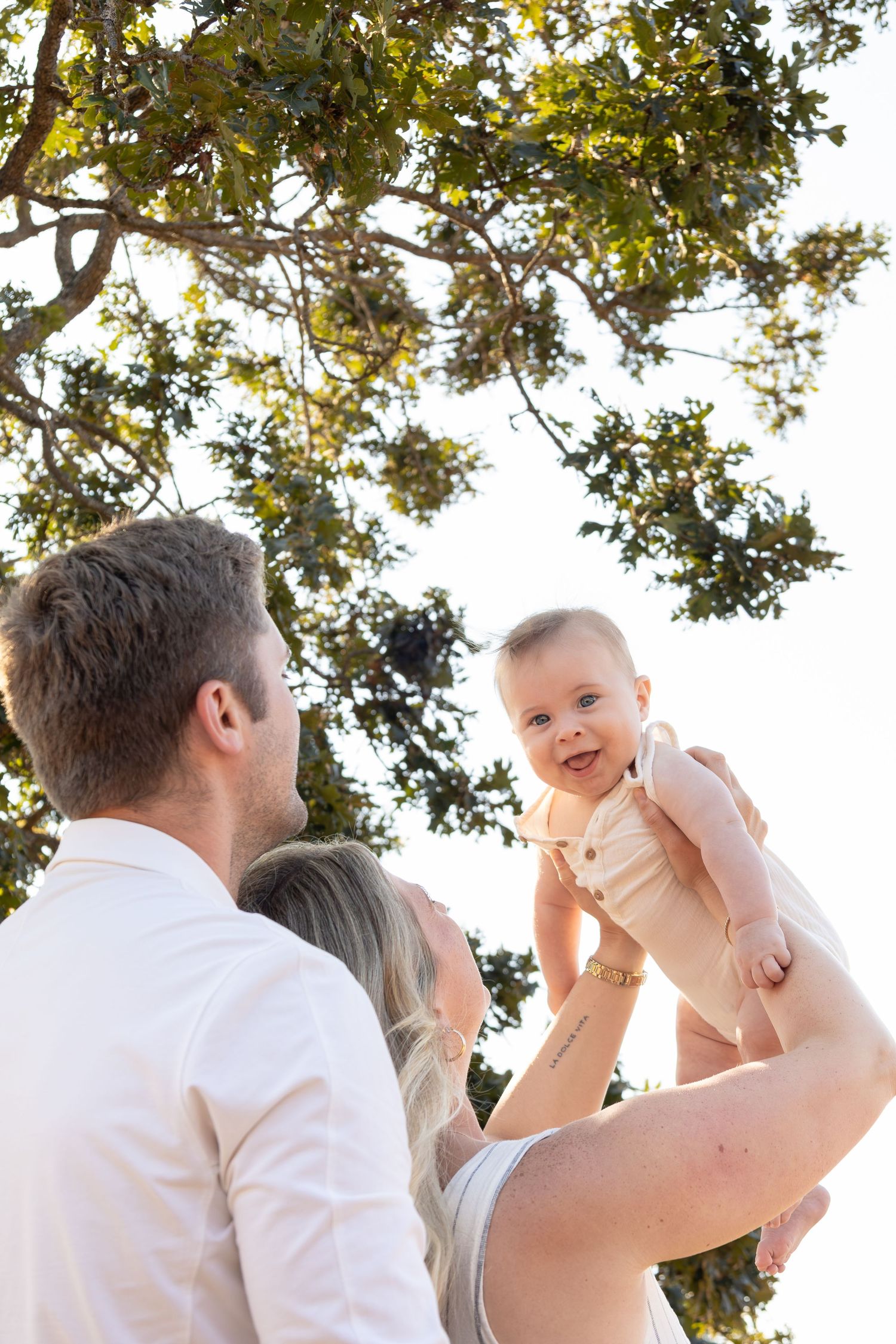 A husband hugs his wife from behind as she holds their baby up in the air. They smile at the baby while the baby smiles at the camera.