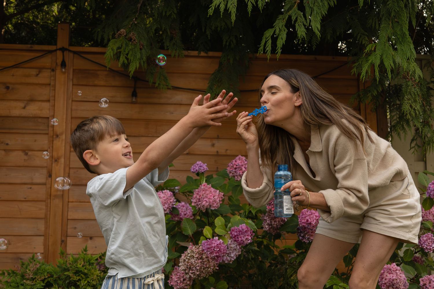 A mom plays in the backyard with her young son. The mom blows bubbles while the son chases after them.