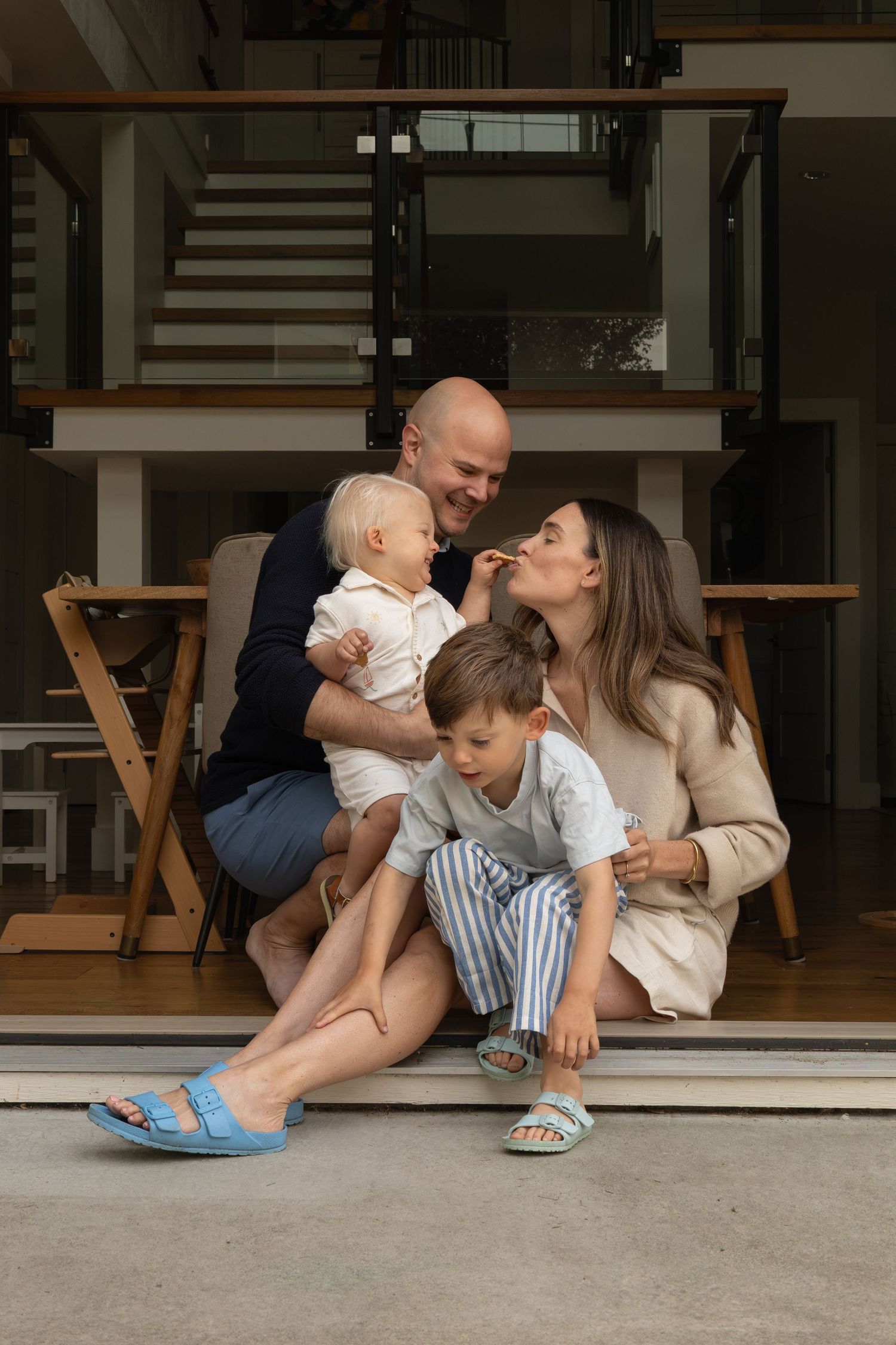 A family with two young boys sits close together in the doorway of their home. The youngest boy feeds his mom an animal cracker.