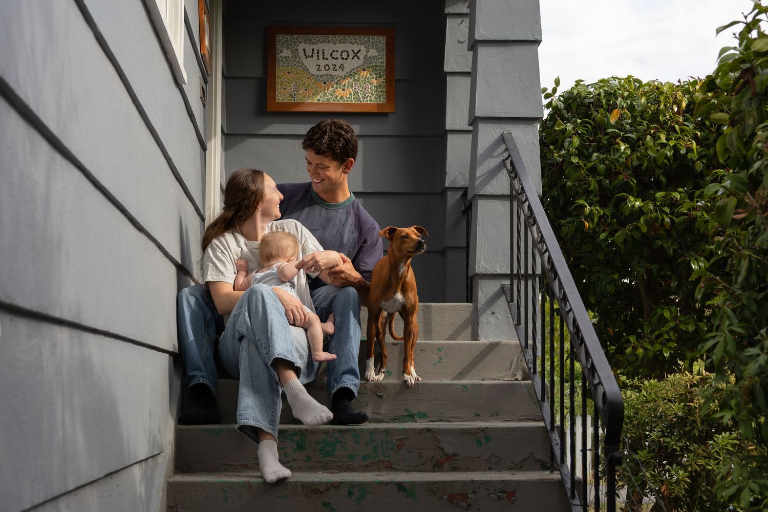 Young parents sit in the sun on the front steps outside their home playing with their baby and their dog.