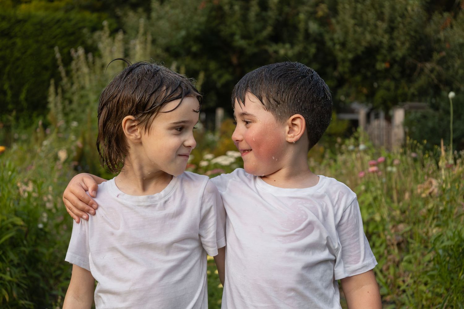 Two young boys stand in a garden with their arms around each others shoulders while they mischievously look at each other.