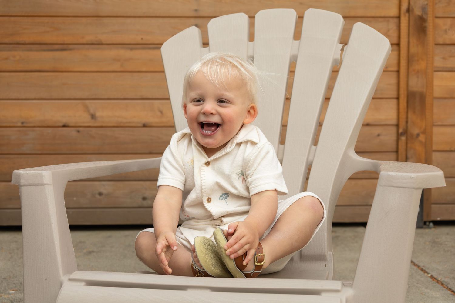 A toddler smiling and laughing while sitting cross legged in a chair in the backyard.