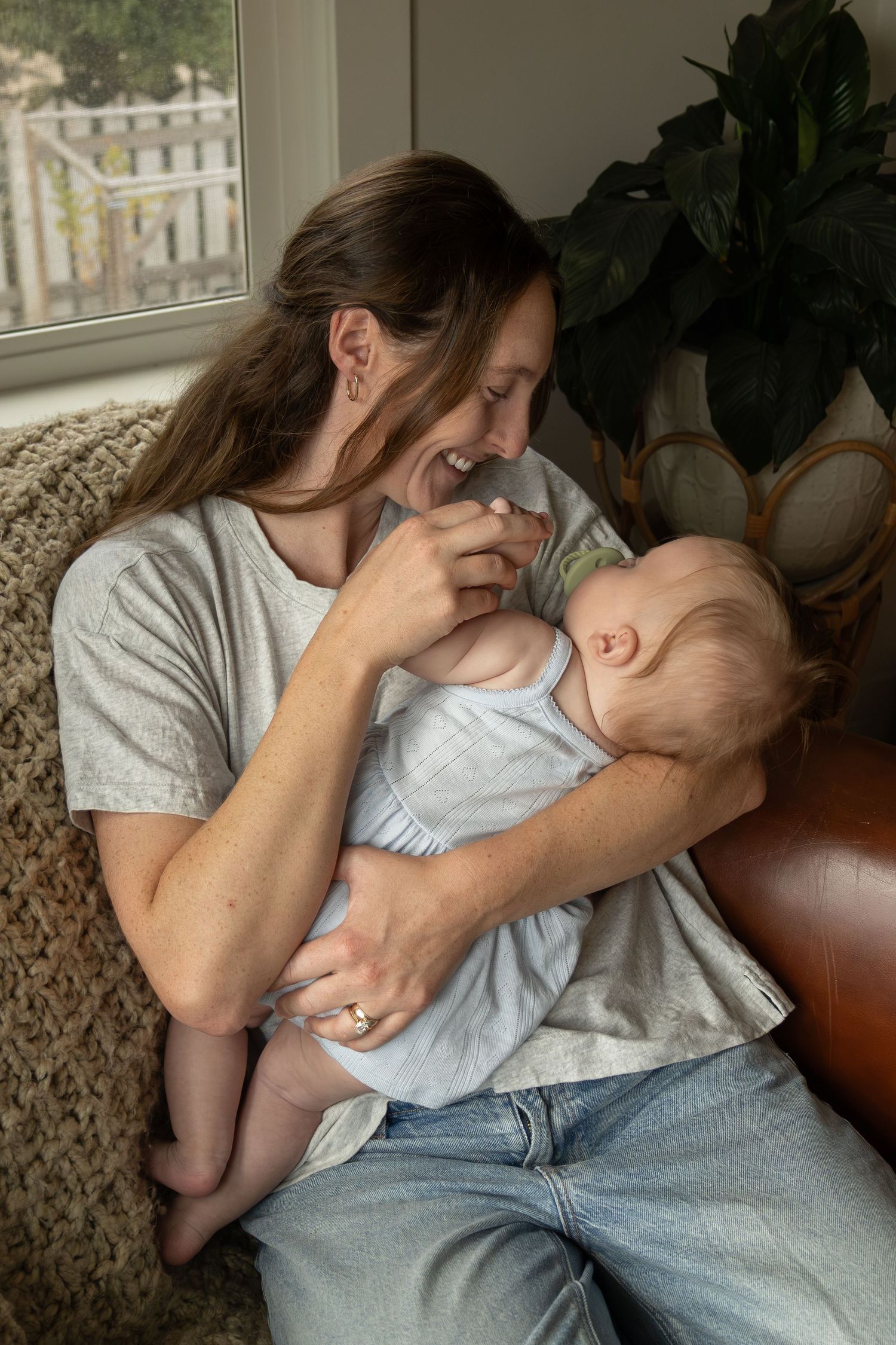 A young mother snuggles her baby and smiles down at her while sitting in a big comfy chair in a living room.