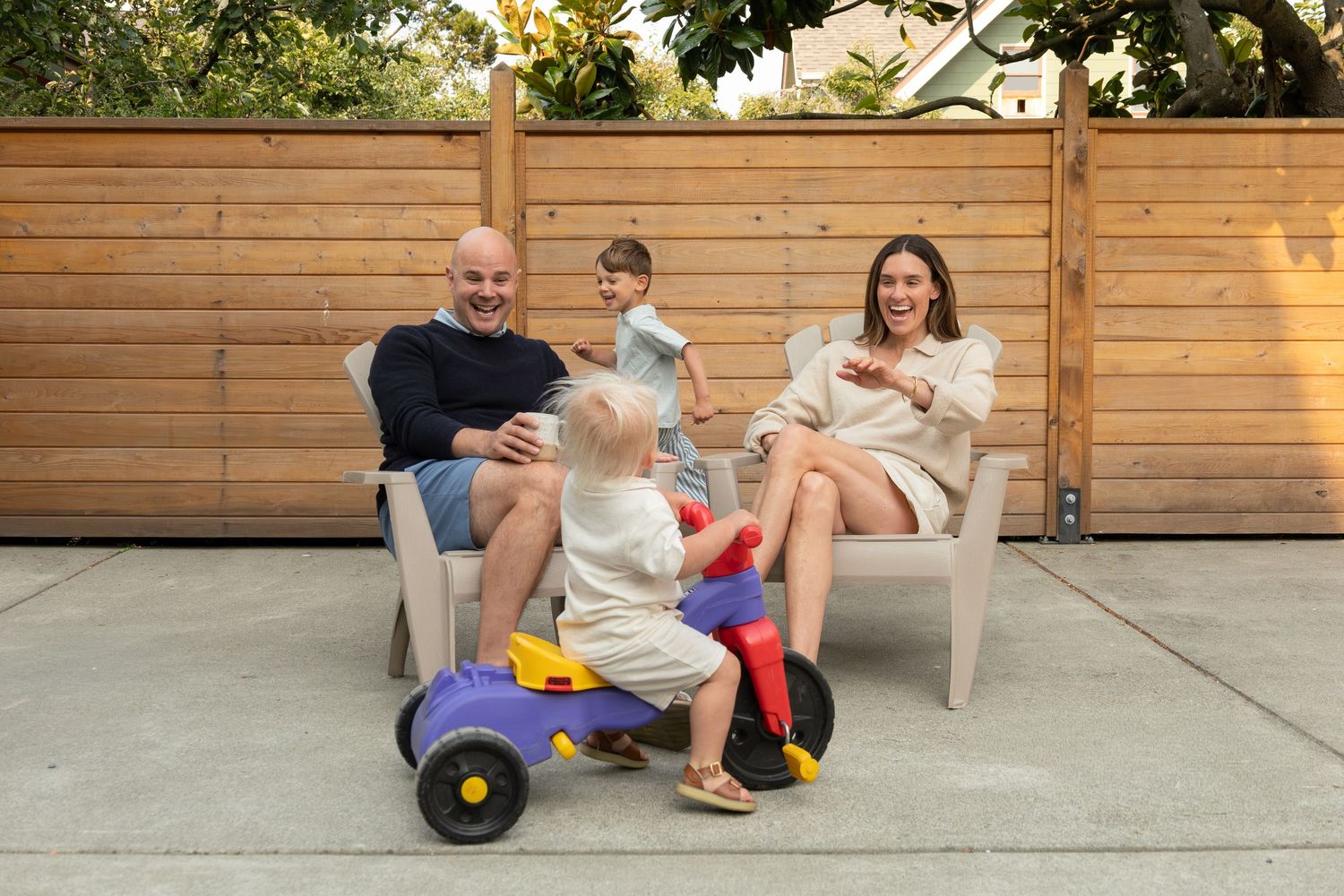 Two parents sit in chairs in the backyard with coffee while their two young sons run around them playing. The parents laugh and wave.