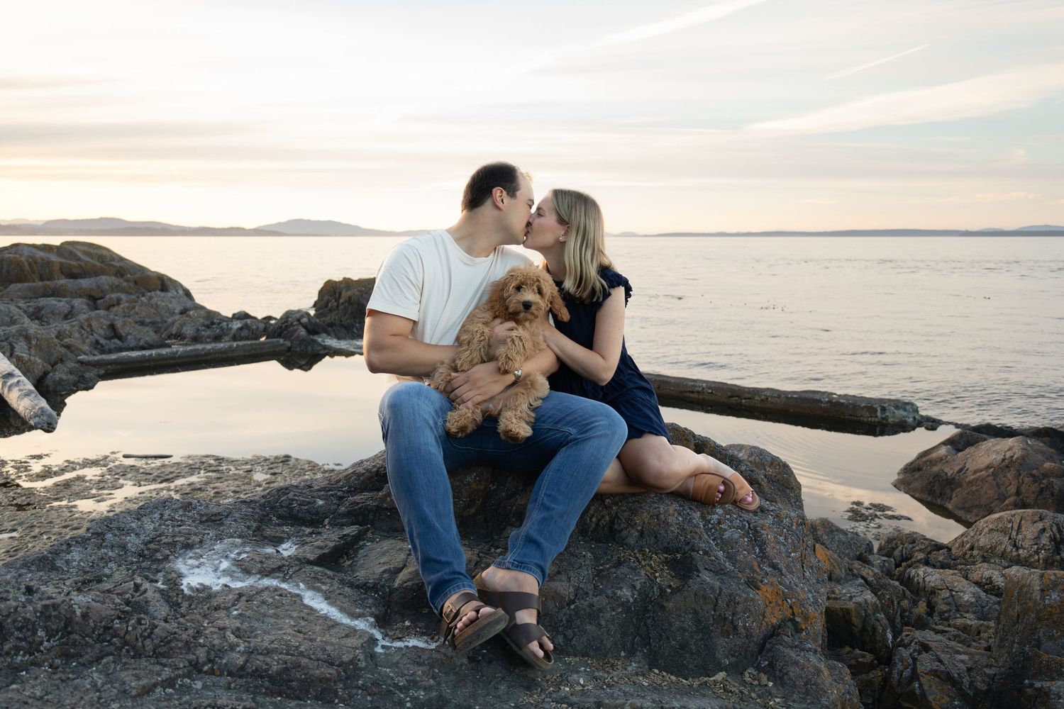 A young couple sits on a large rock beside the ocean at sunset. They kiss while holding a new puppy in their laps.