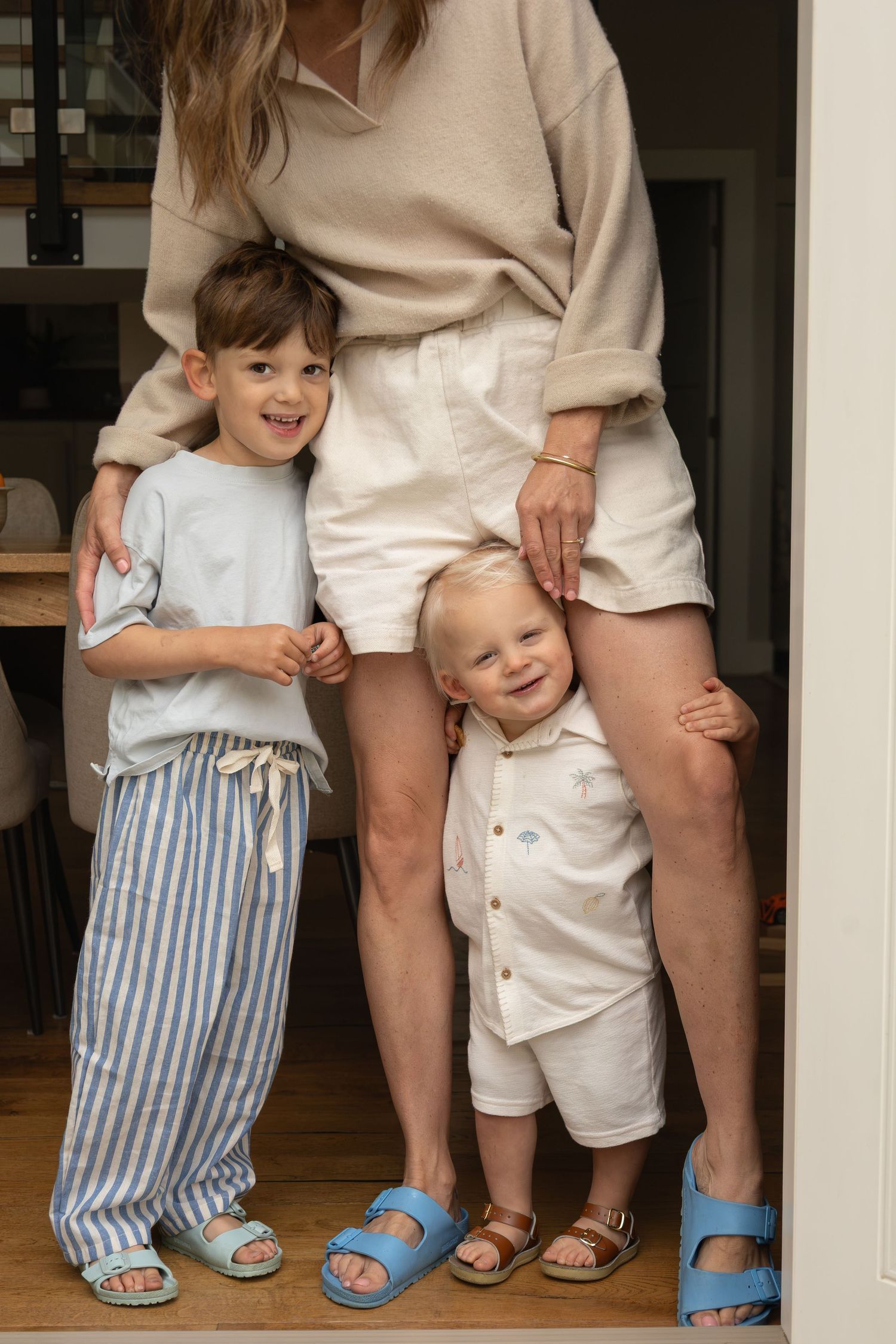A mom stands inside the front door of her home with her two young sons. One is hugging her from the side, the other stands between her legs.