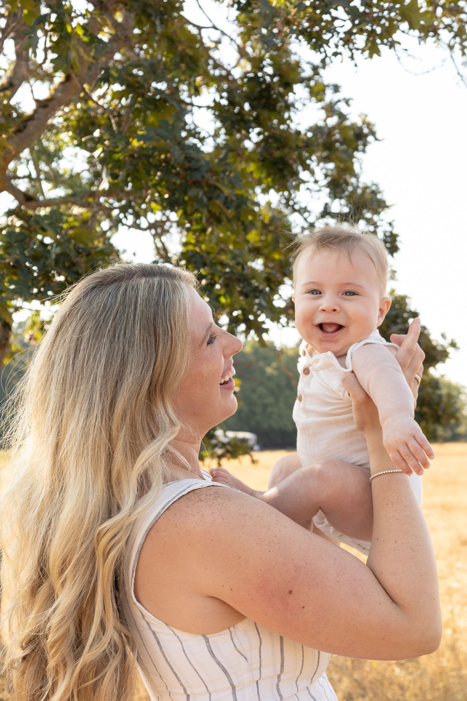 A mom smiles at her baby while holding him up in the air. The baby laughs at the camera.