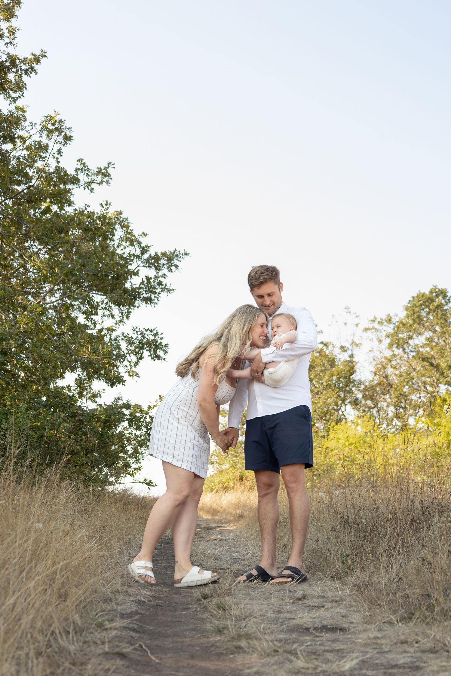 Young parents stand on a pathway in a field holding their new baby. The couple holds hands while the mom leans down to kiss the baby.