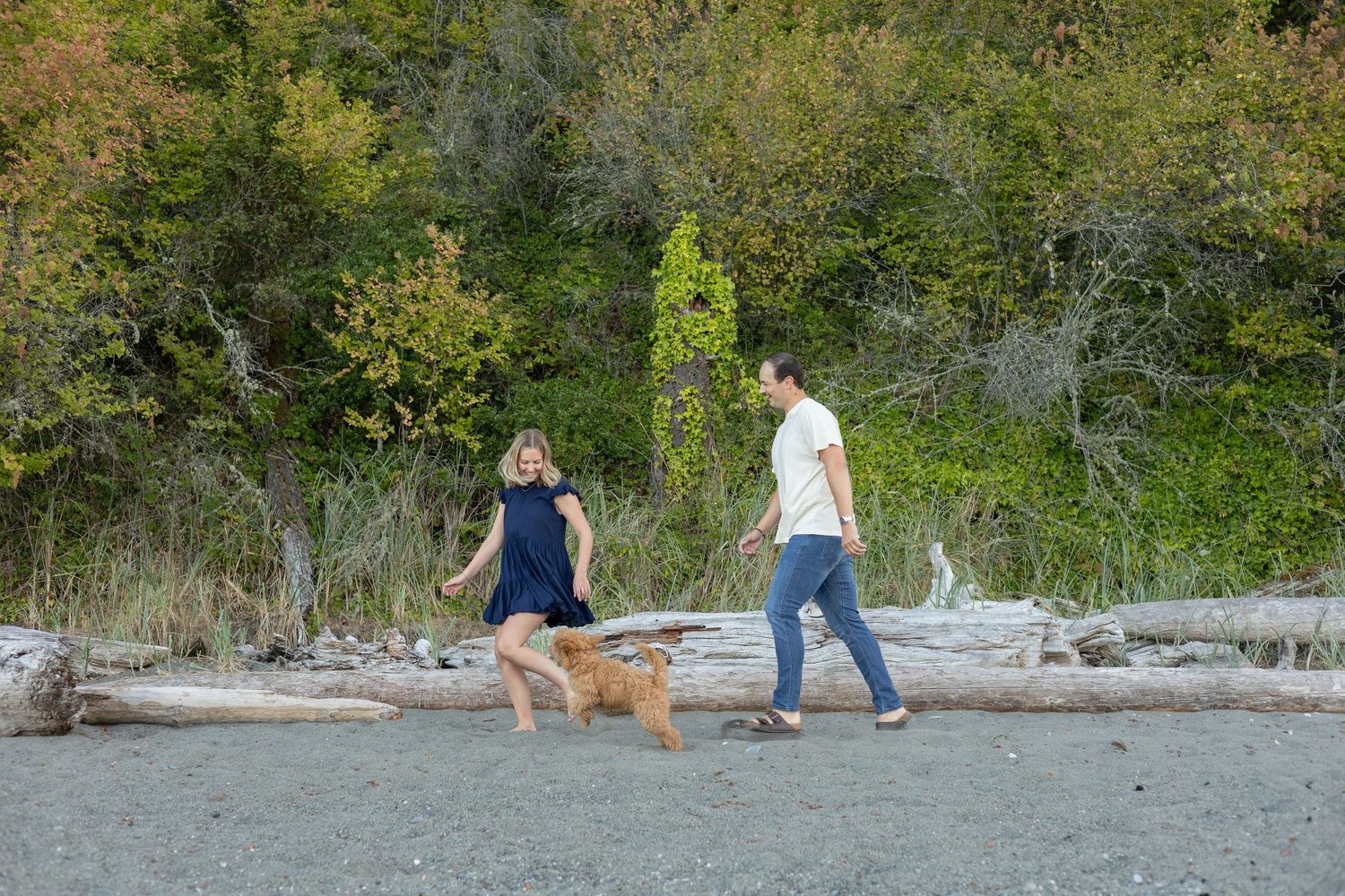 A young couple runs along the beach with their new puppy chasing after them. The women looks down and smiles at the puppy.