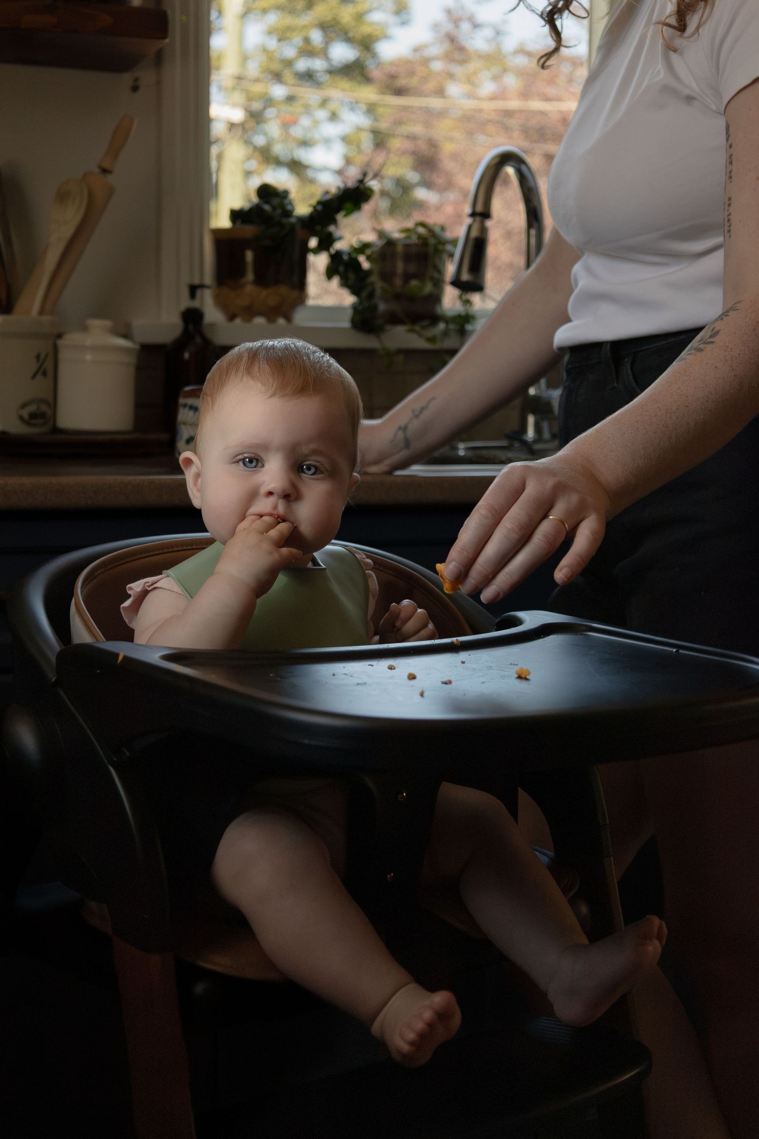 A baby sits in a high chair eating food while looking at the camera. The mom stands beside trying to help.