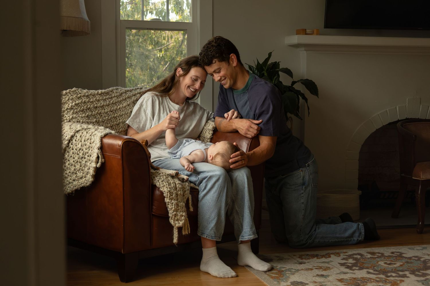 A mom sits in a chair with a baby laying in his lap. The dad kneels beside them on the floor. Both parents smile at the baby.