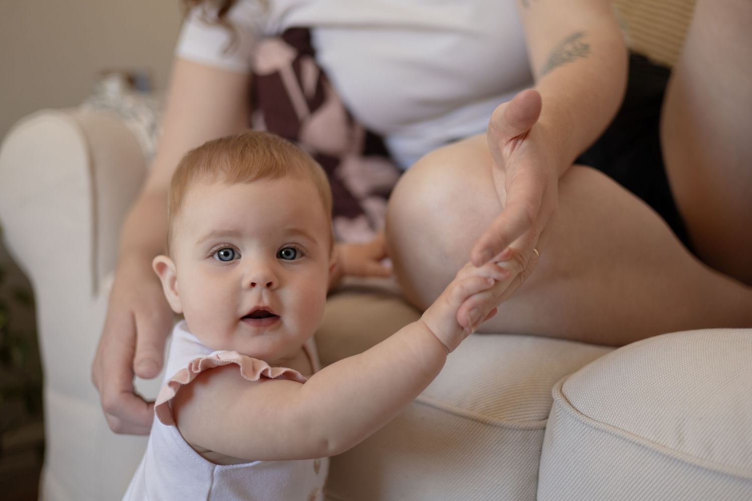 A baby stands up and looks at the camera while using their mom's hand to help stand up.