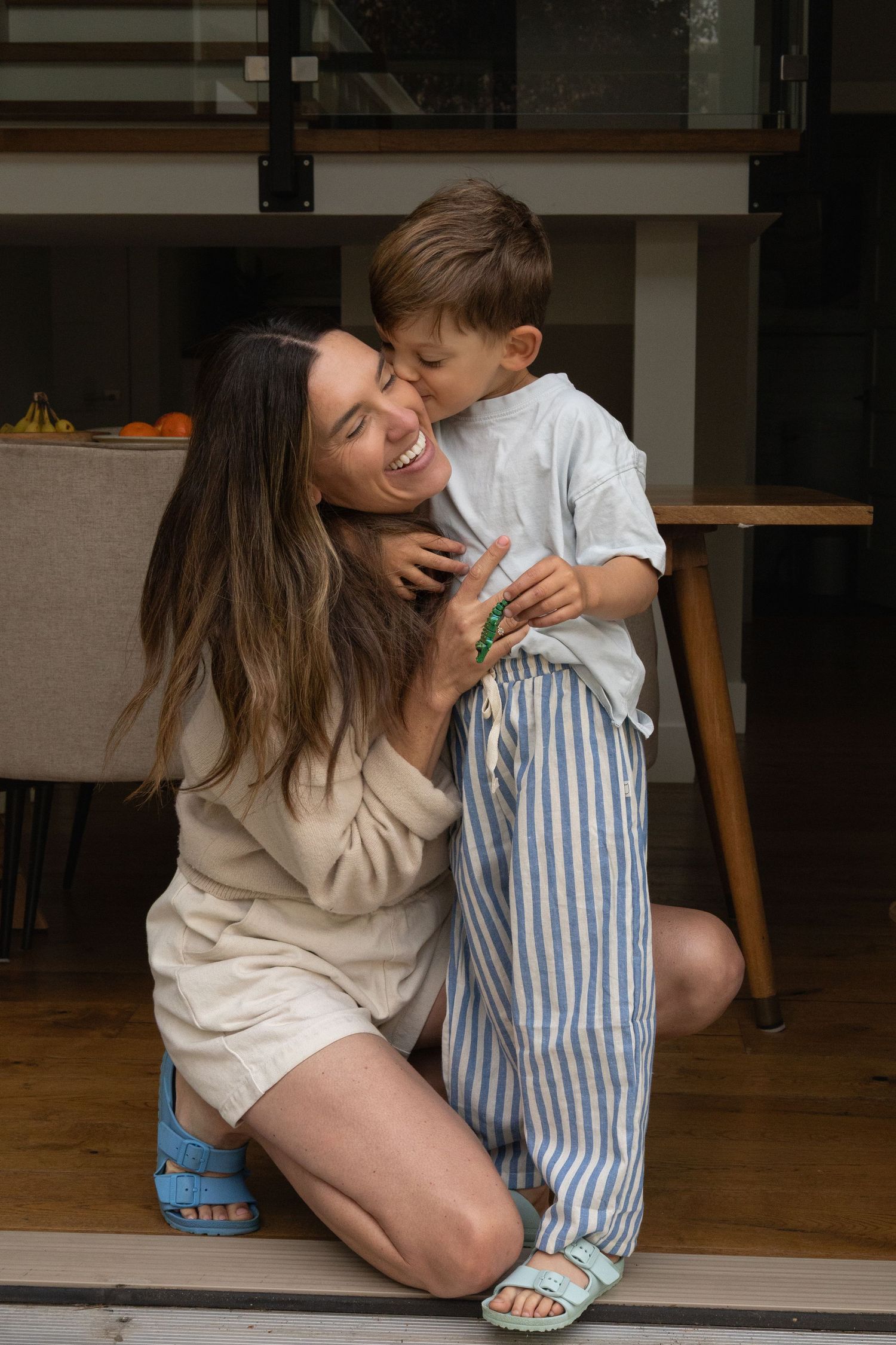 A mom kneels down to hug her young son in their front doorway. The boy kisses the mom on the cheek while she closes her eyes and smiles.