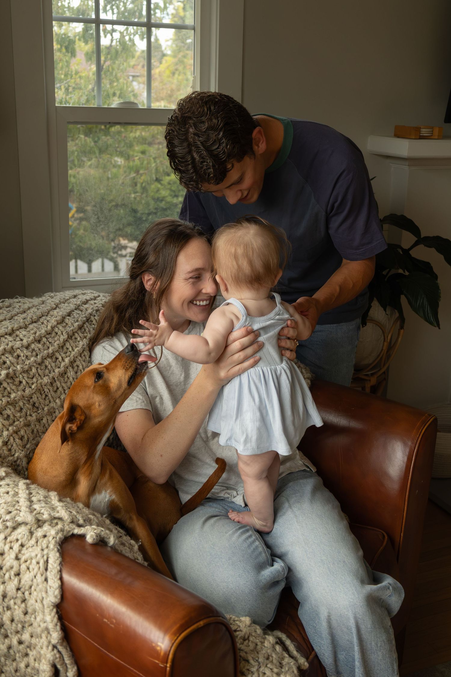 A young mom sits in a chair in a living room with a dog while holding a baby who is standing on her lap. The dad stands behind watching.