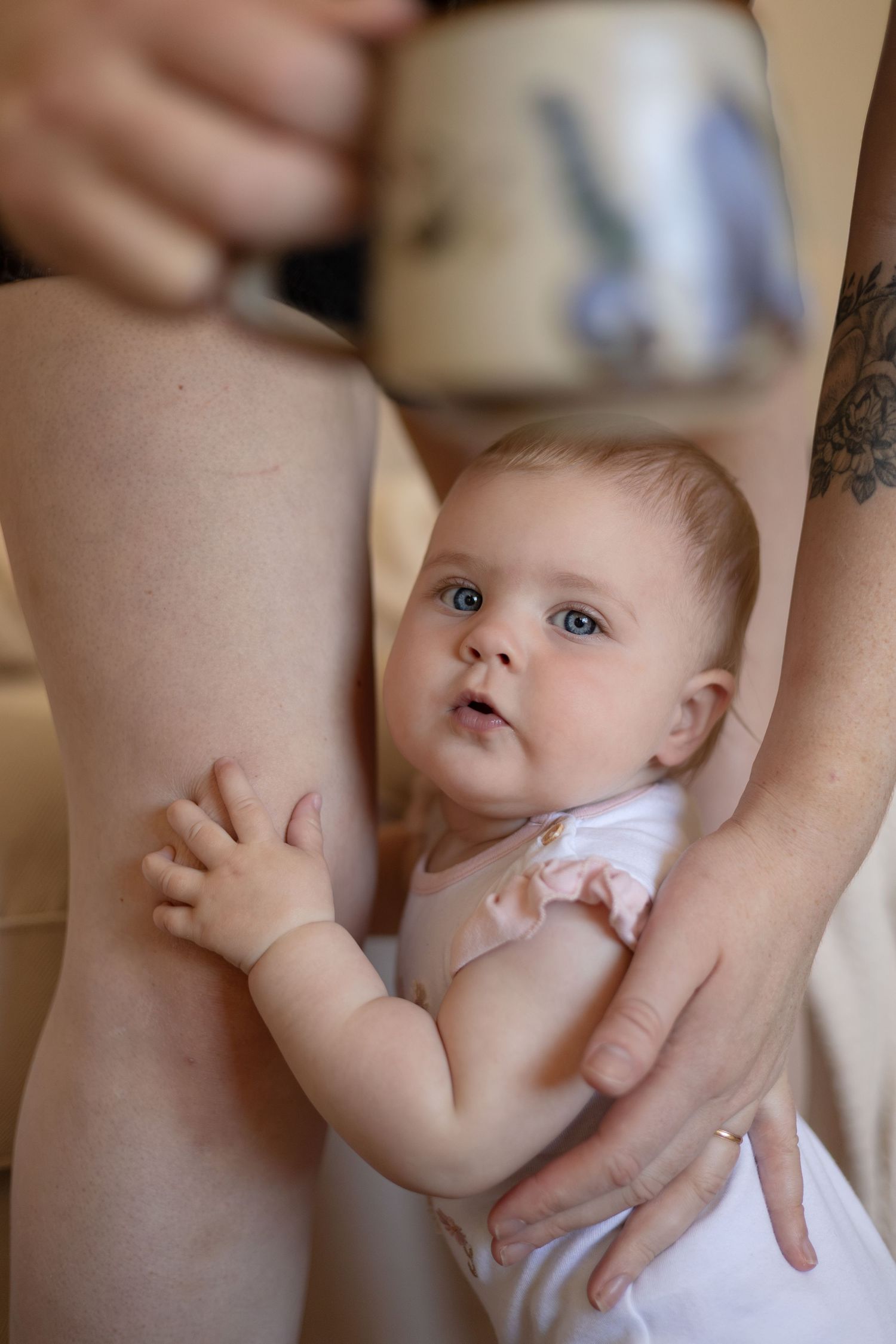 A baby stands up and looks at the camera while holding onto their mom's legs to help balance.