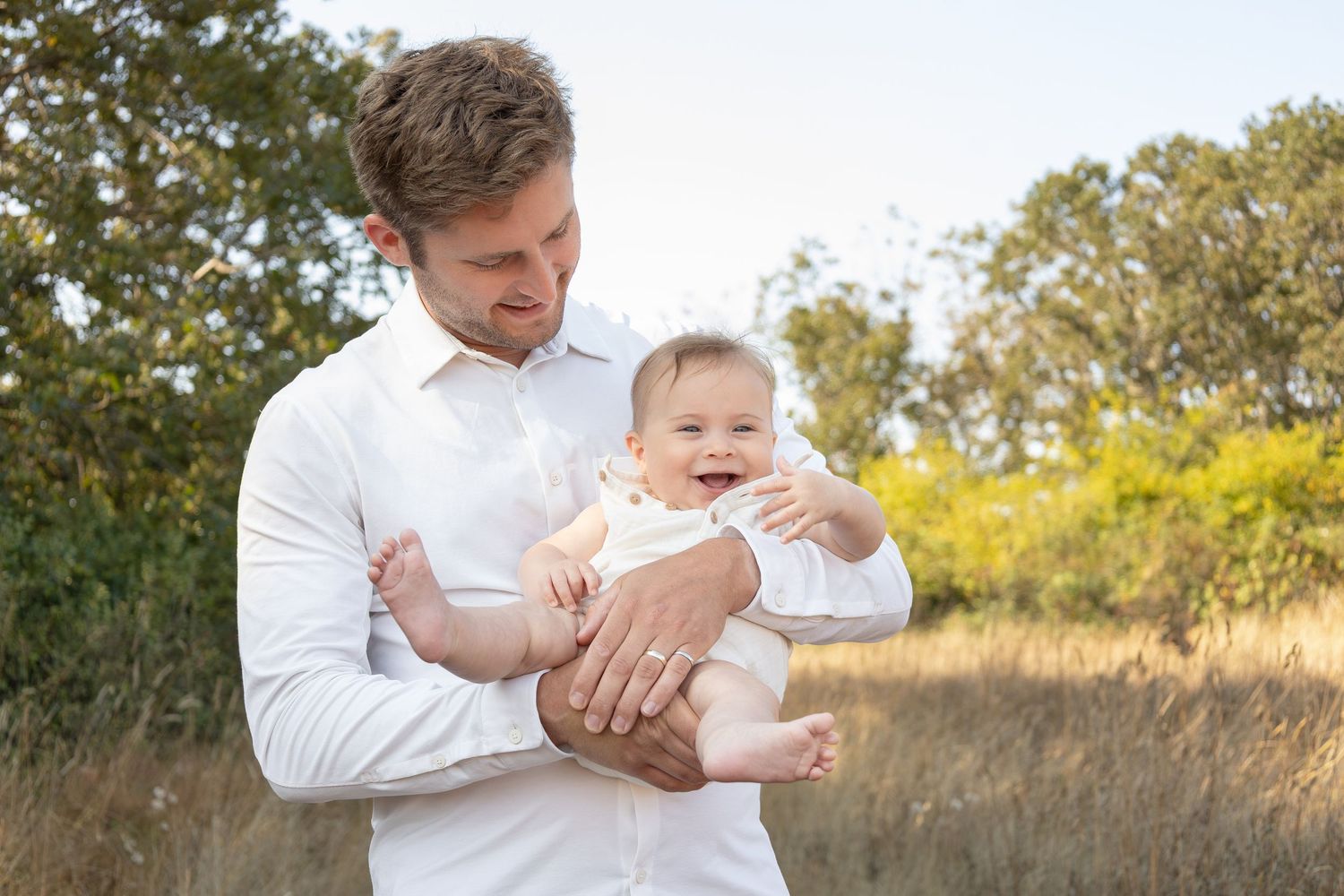 A dad stands in a field holding his baby. He looks down at the baby and smiles while the baby smiles and looks out towards the camera.