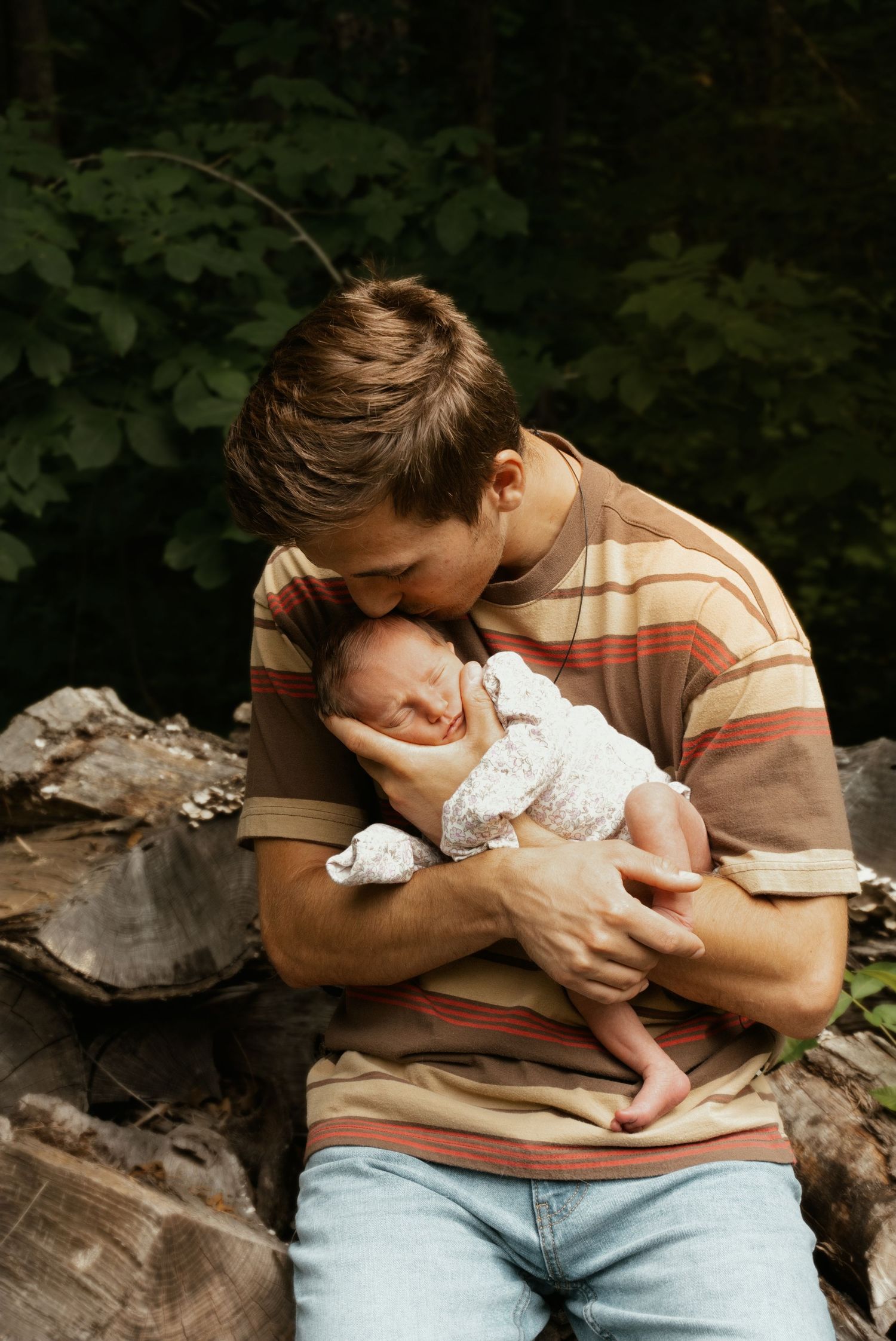 New father in striped shirt tenderly kisses his sleeping newborn baby while sitting outdoors on logs.