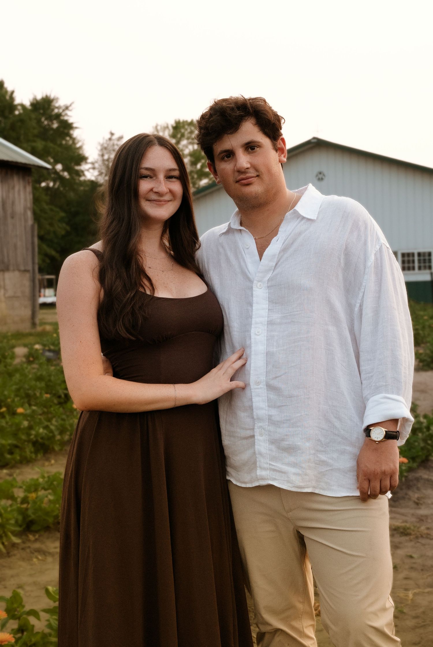 Couple posing together near a farm barn, woman in brown maxi dress and man in white linen shirt.