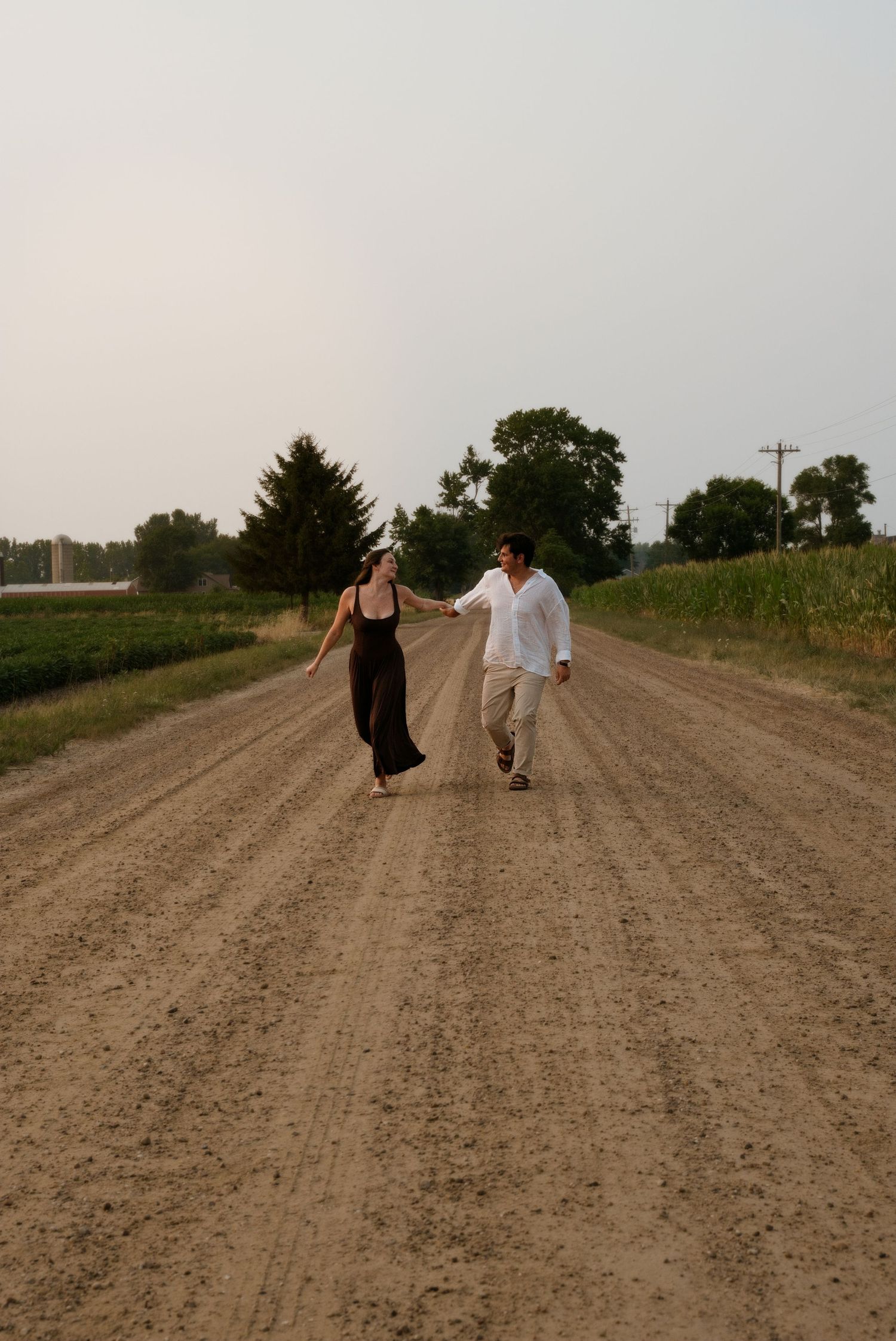 A couple holds hands and walks together on a rural dirt road surrounded by green fields and trees at dusk.
