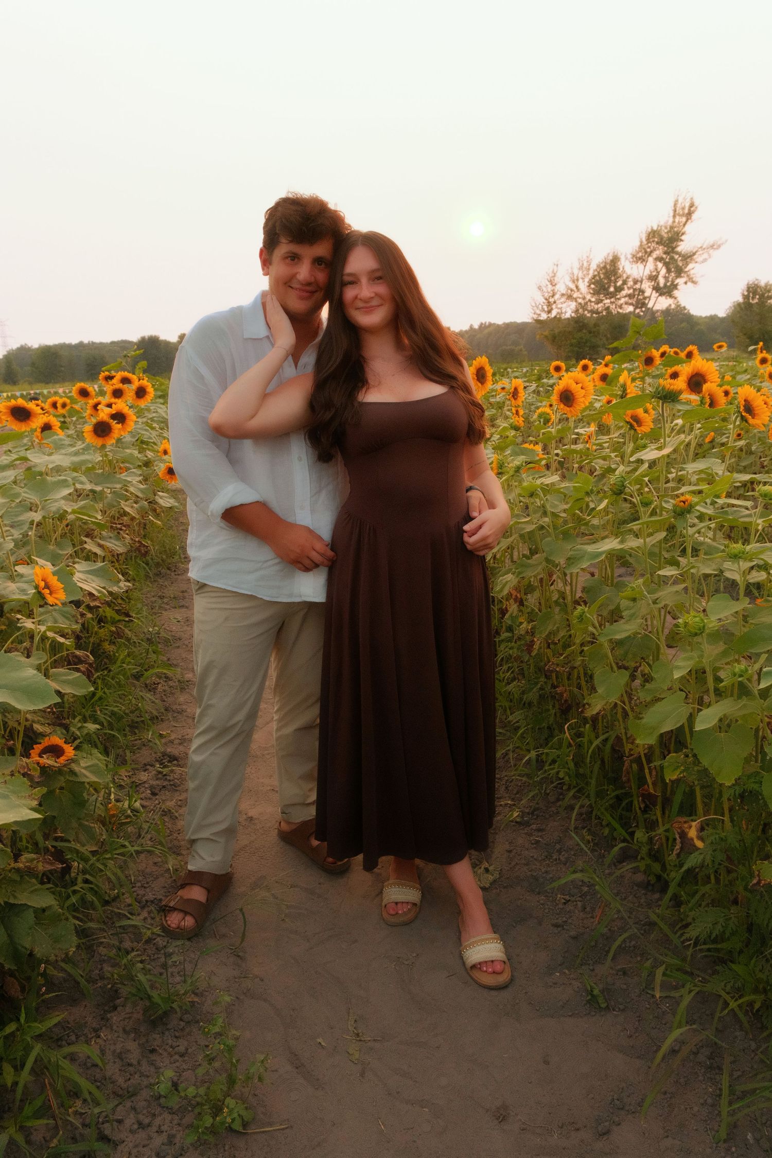 Smiling couple posing together in a sunflower field during a warm golden sunset.