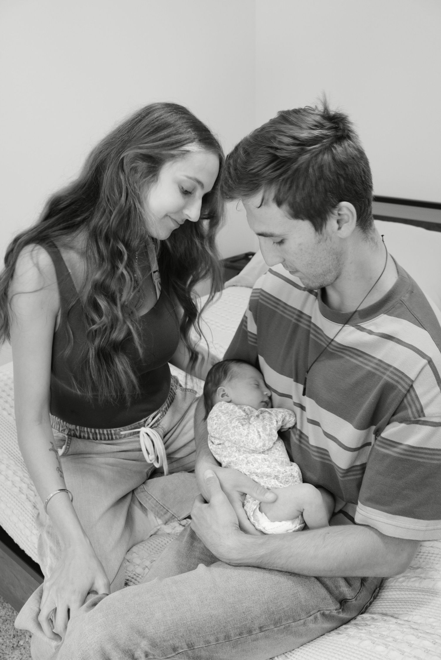 Black and white photo of young parents sitting on bed, lovingly gazing at their newborn baby.