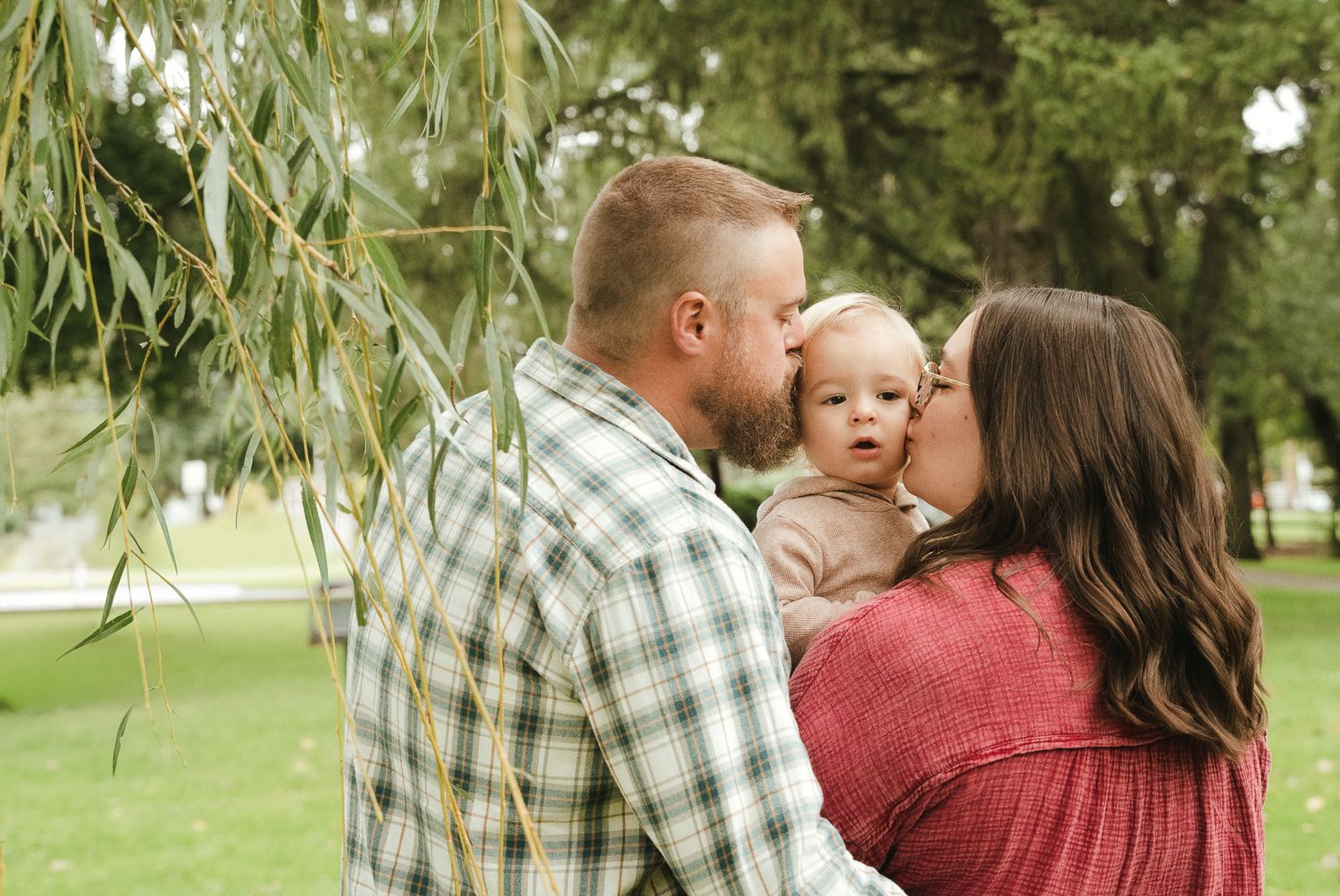 A man in a plaid shirt and a woman in a red top kiss a baby between them in a sunny park beside a weeping willow tree.