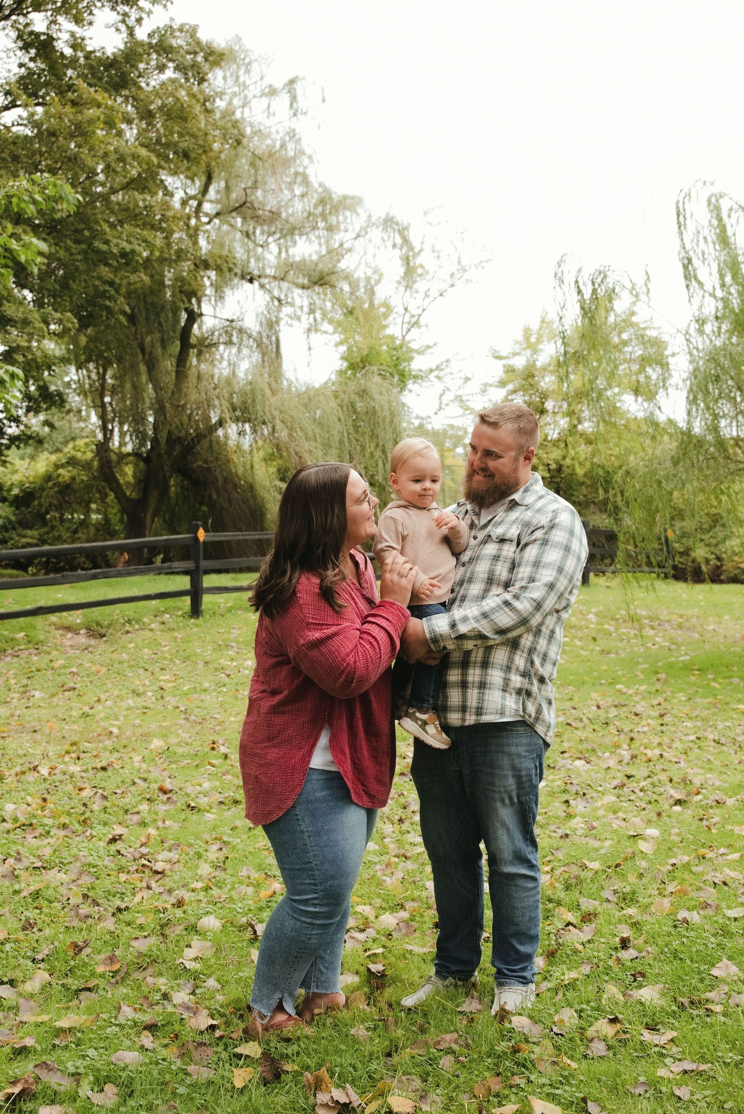A smiling family of three poses outdoors on a leaf-covered lawn as a woman and bearded man hold their toddler near willow trees.