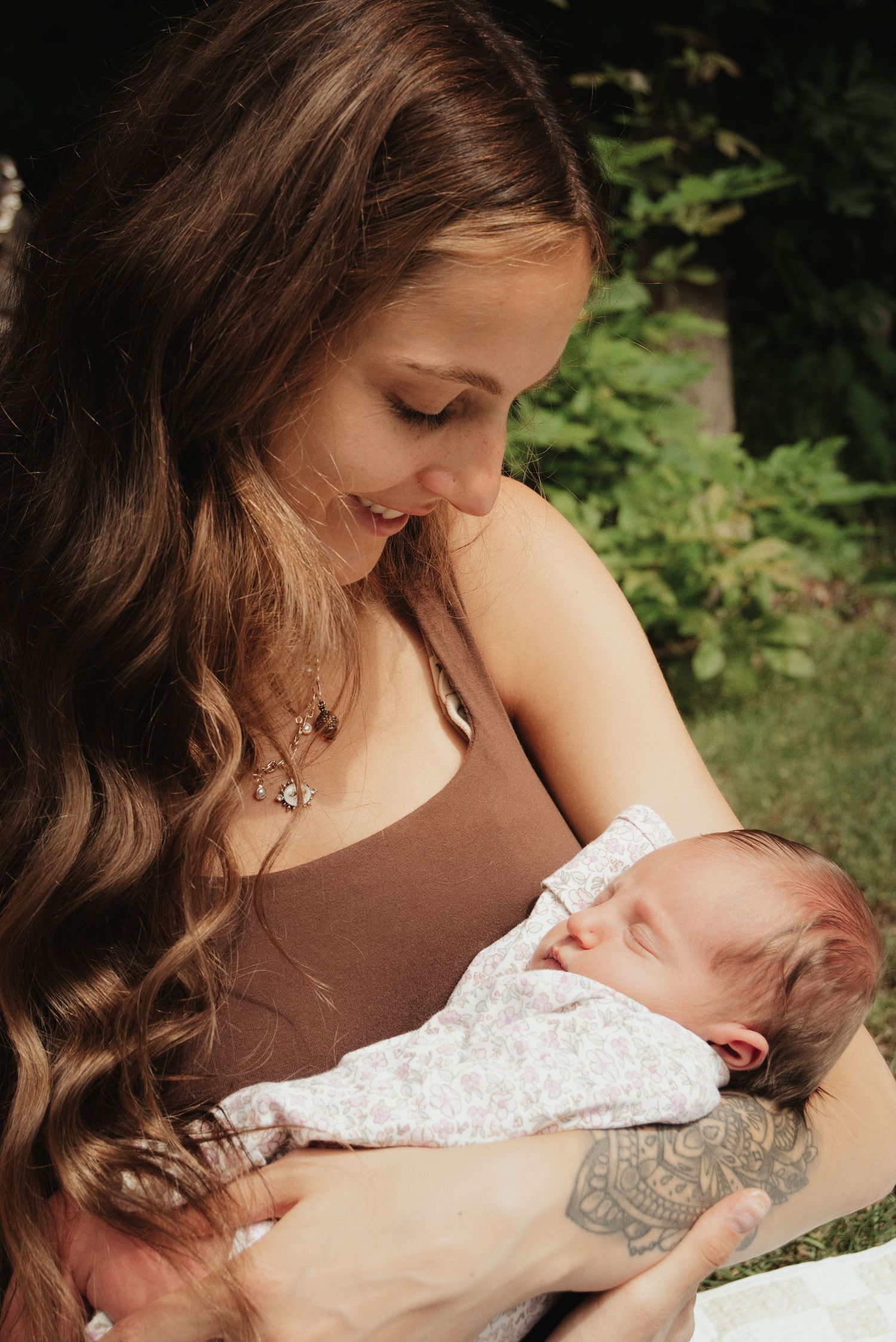 Young brunette mother smiling down at her sleeping newborn baby cradled in her arms outdoors in garden.