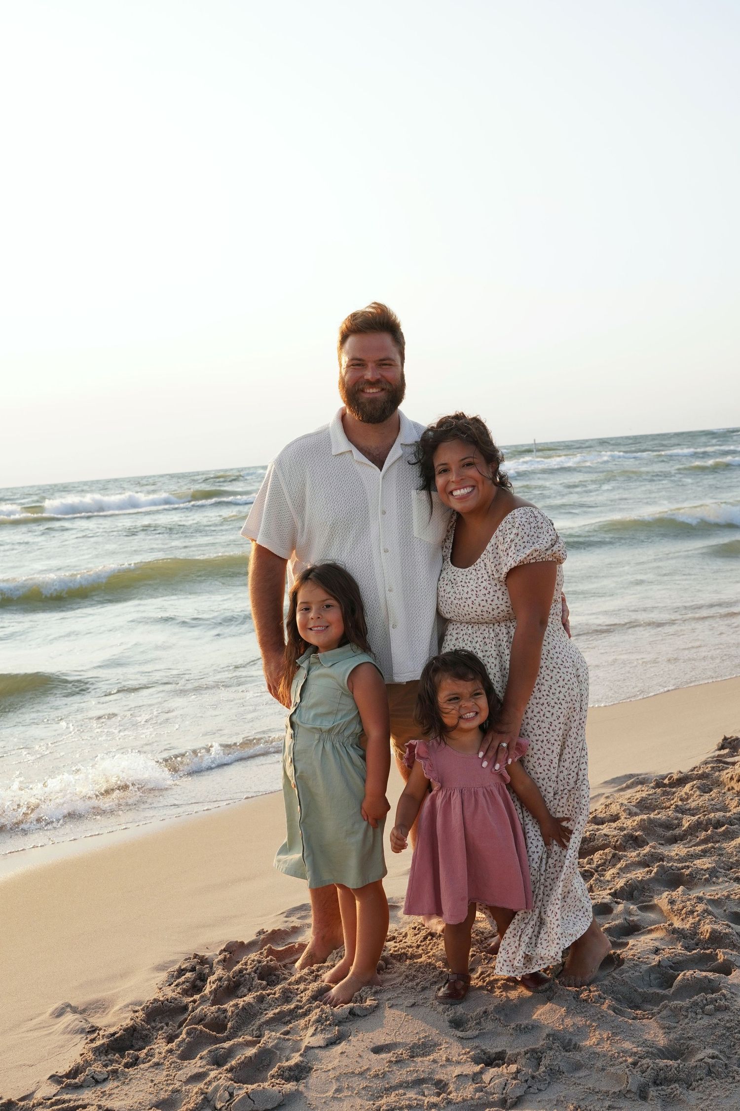 Family of four smiling together on a sandy beach with ocean waves in the background at sunset.