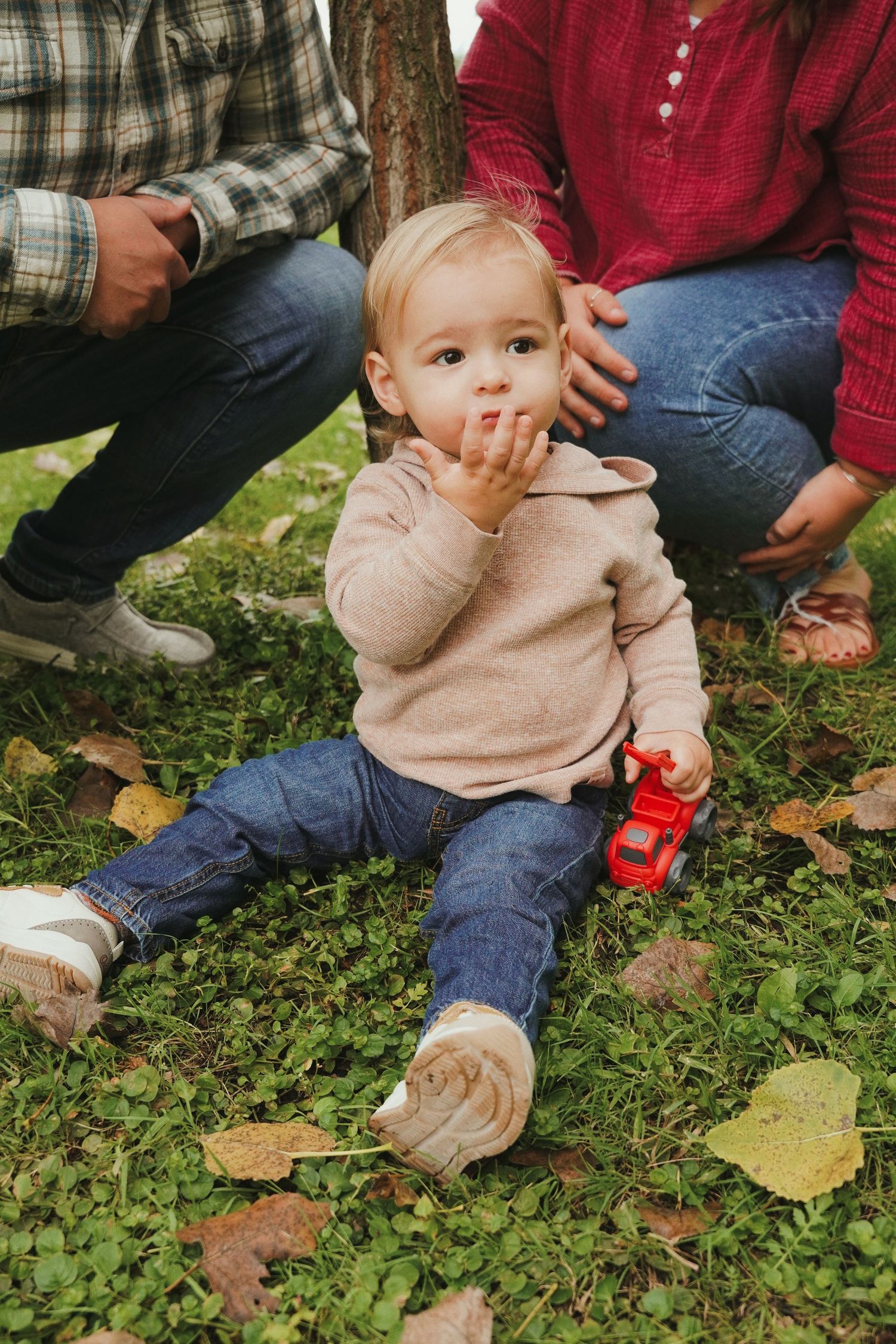 A toddler in a beige hoodie and jeans sits on grassy ground with fallen leaves, holding a red toy truck while two adults crouch nearby.