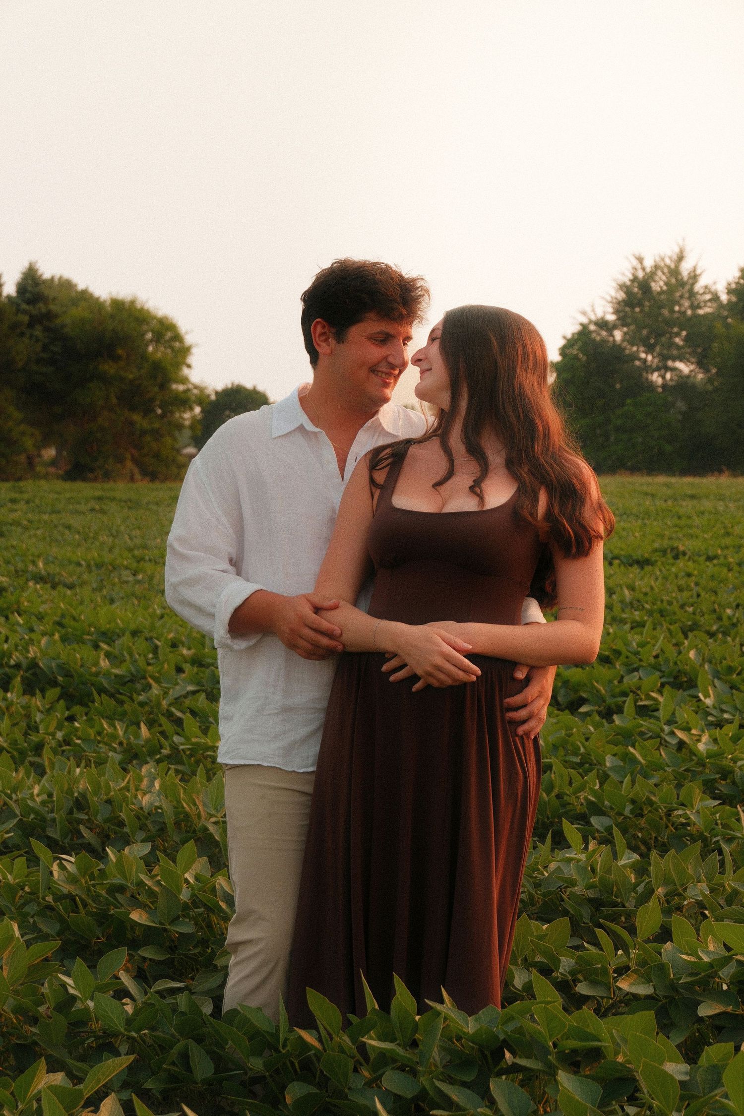 Expecting couple sharing a tender moment in a green field at sunset, woman in brown maternity dress.
