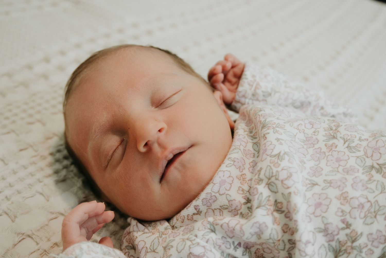 Peaceful newborn baby sleeping soundly wrapped in a soft floral swaddle blanket on a white textured surface.