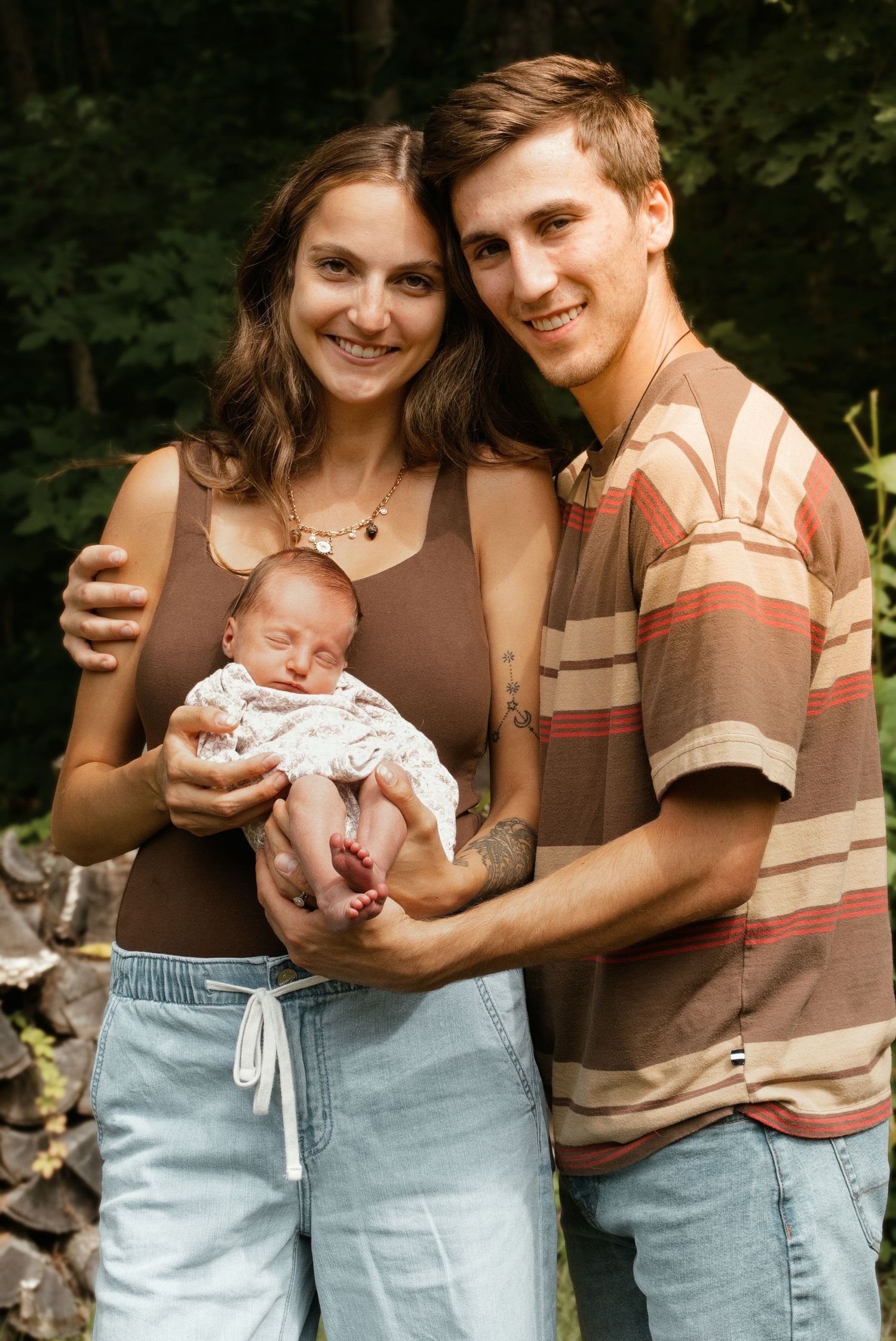 Happy young couple smiling and holding their newborn baby together outdoors in a lush green setting.