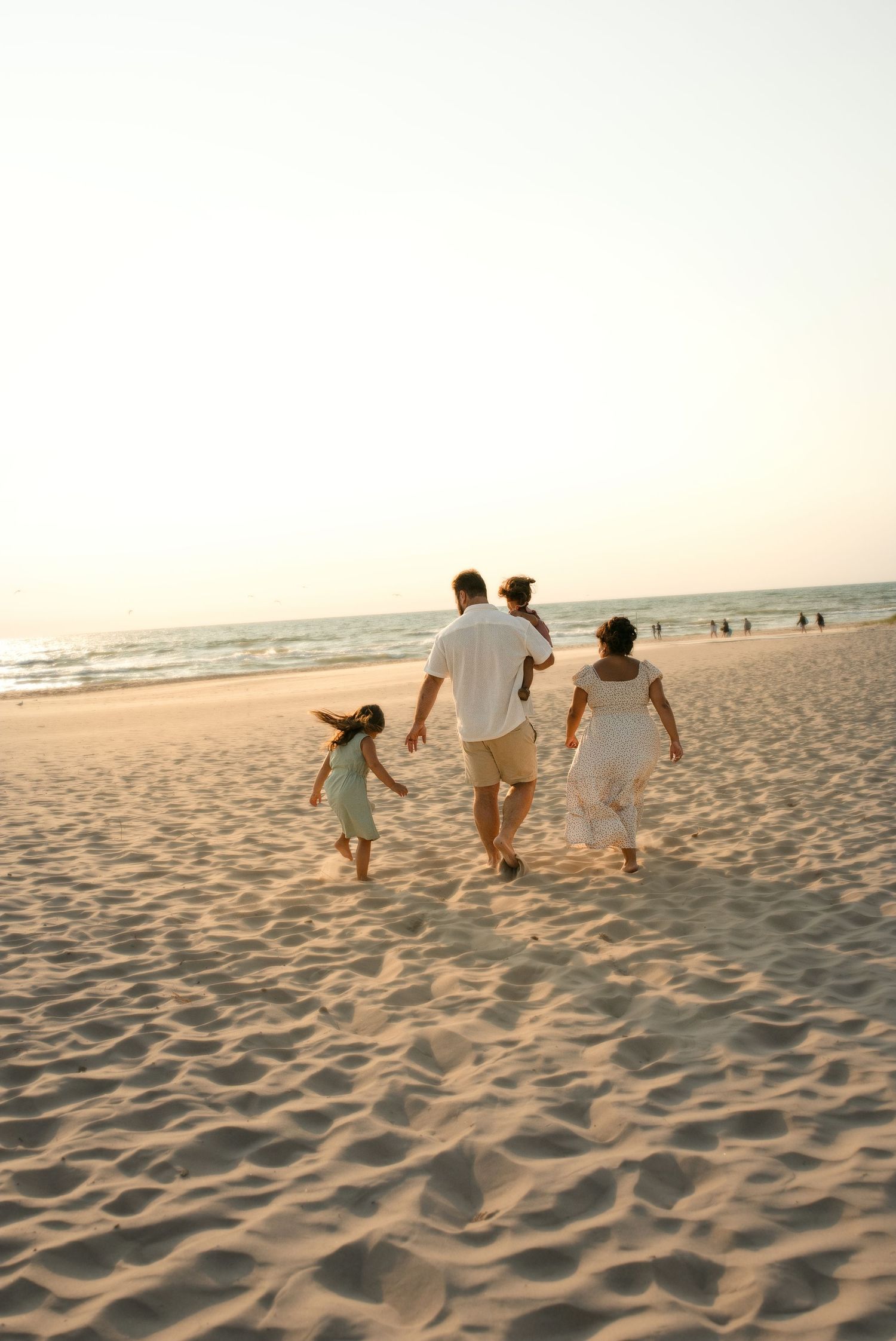 Family of four walking hand-in-hand on a sandy beach at sunset, dressed in white clothing.