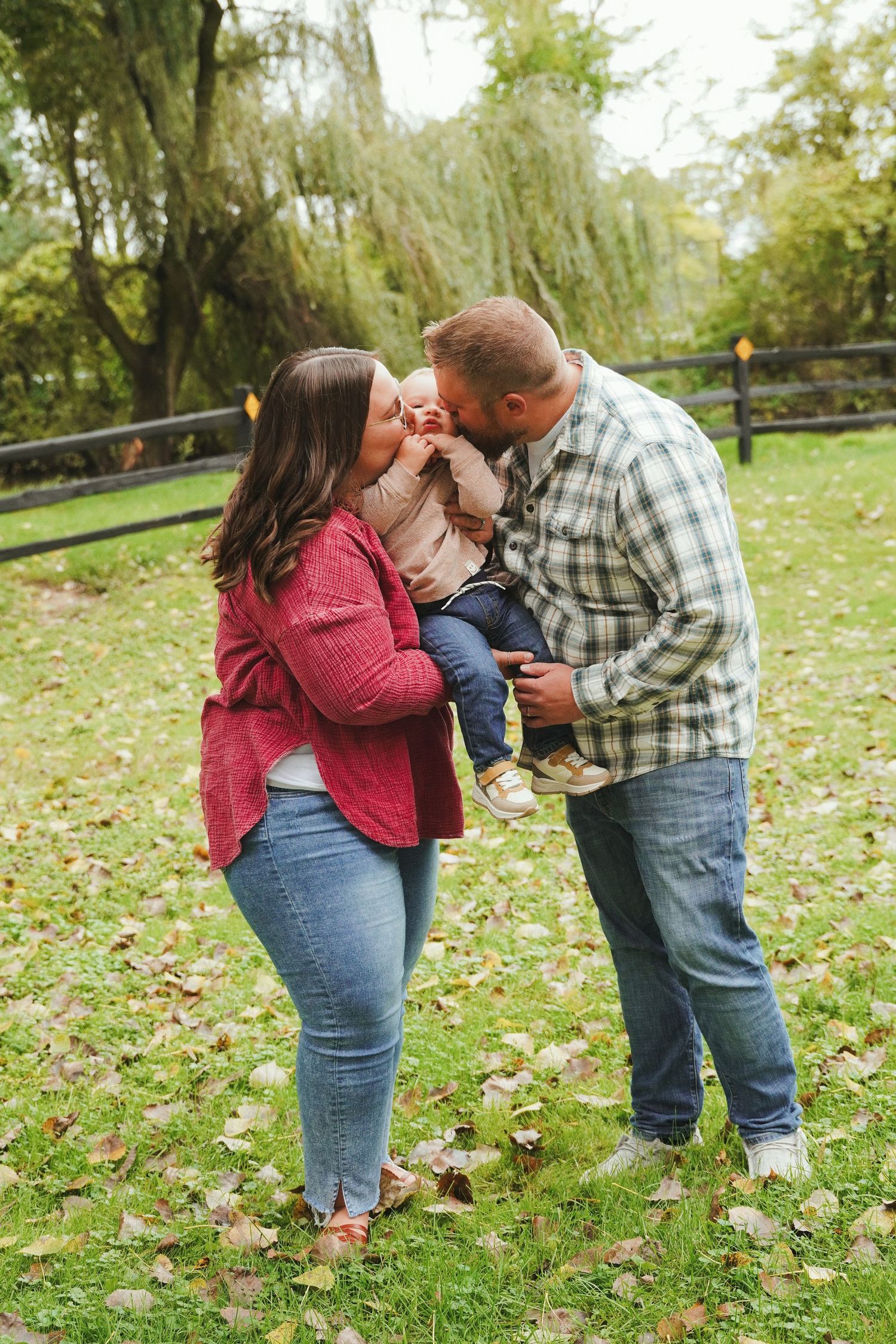 Smiling couple kissing their toddler's cheeks during an outdoor family photo session on a grassy lawn with fall leaves and willow trees.