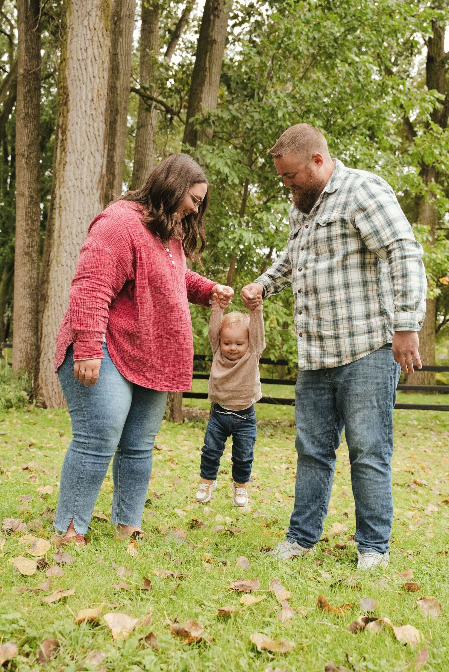 Smiling parents hold a toddler's hands and lift him off the ground while standing on a grassy, leaf-covered lawn surrounded by tall trees.