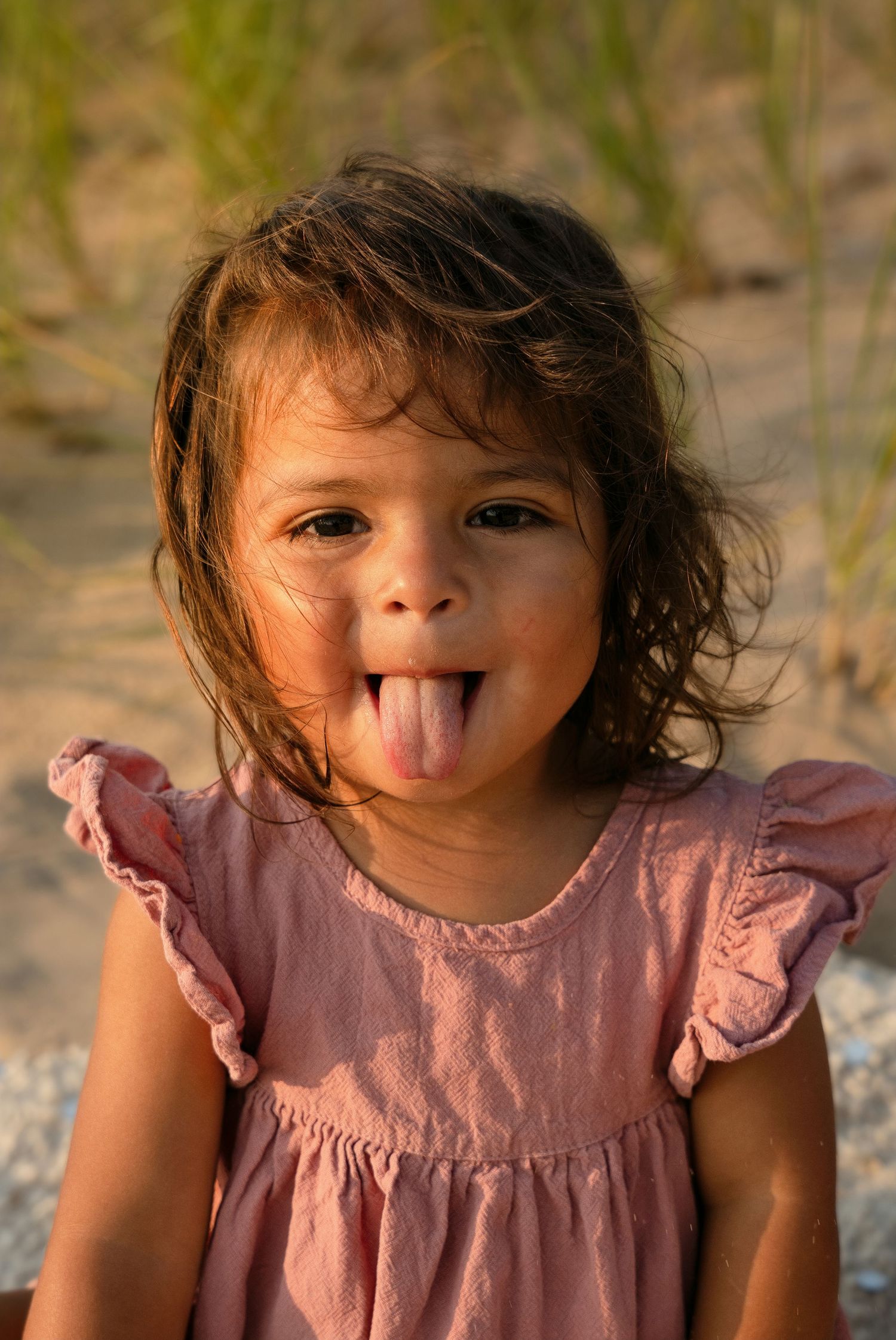 Young girl in pink dress sticking out her tongue playfully on a sandy beach at golden hour.