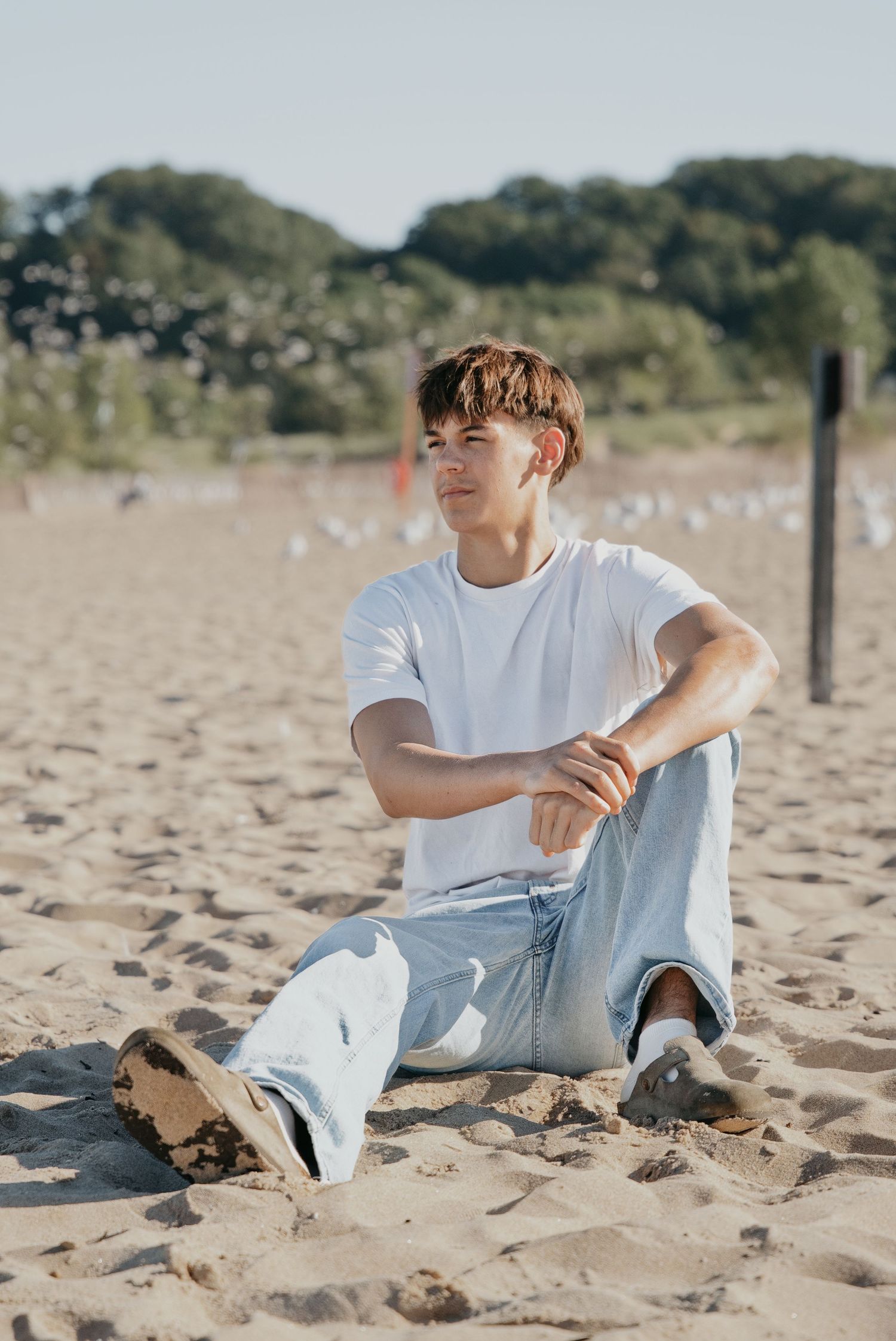 Young man in white t-shirt and jeans sitting relaxed on sandy beach with trees in background on sunny day.