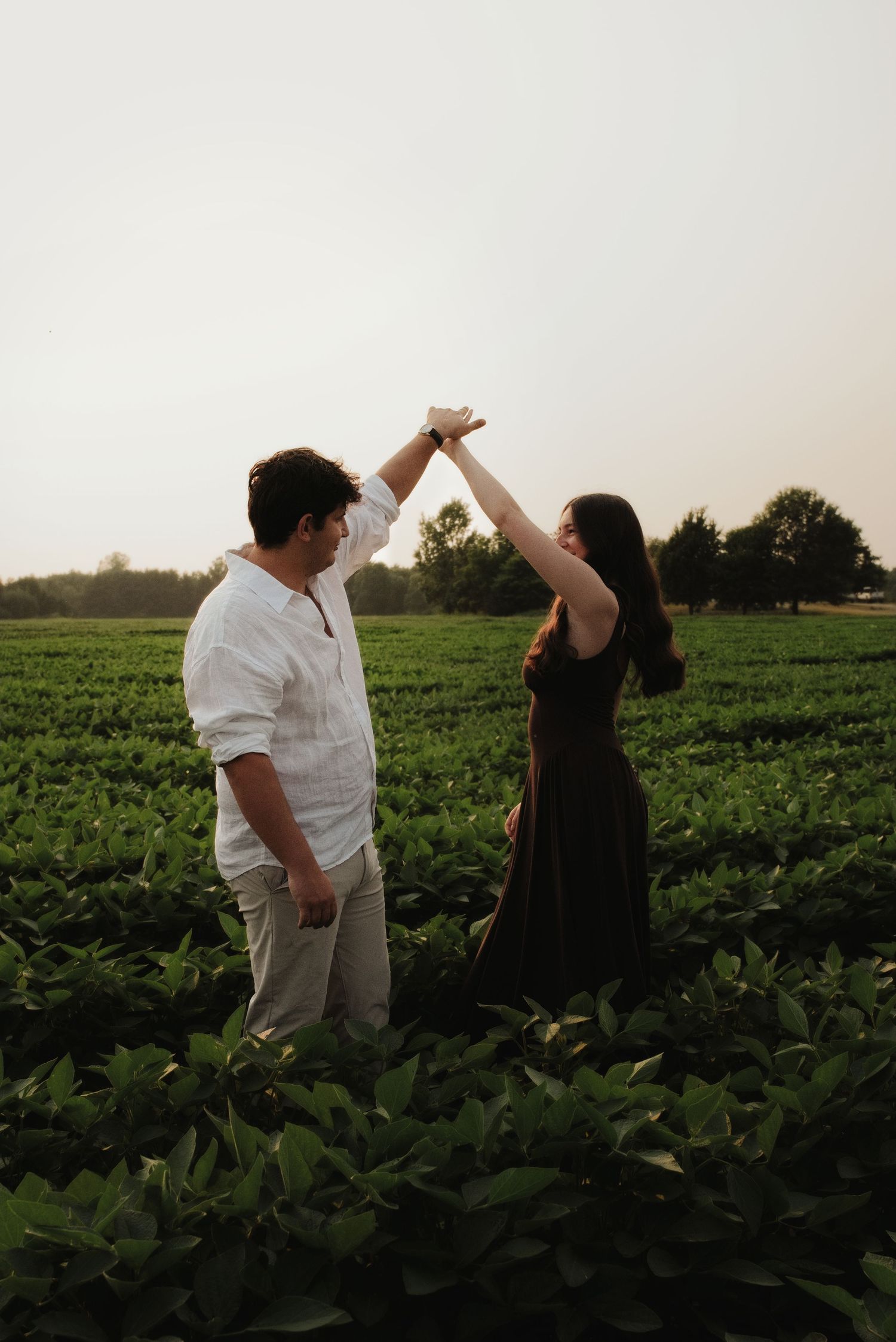 Couple dancing in a lush green field at sunset, man in white shirt raises woman's hand in a romantic twirl.