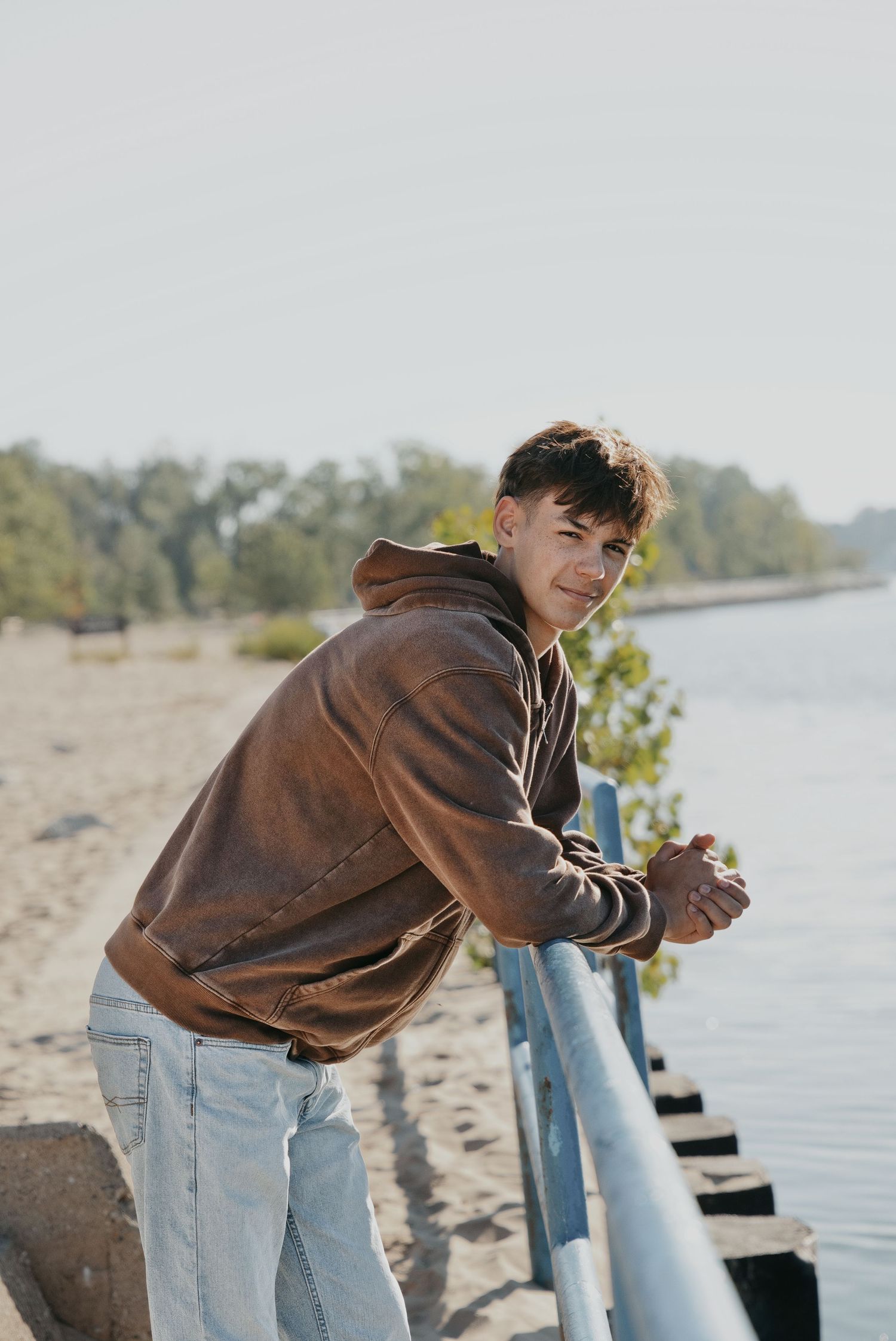 Young man in brown hoodie and jeans leaning on railing by sandy beach shoreline on a sunny day.