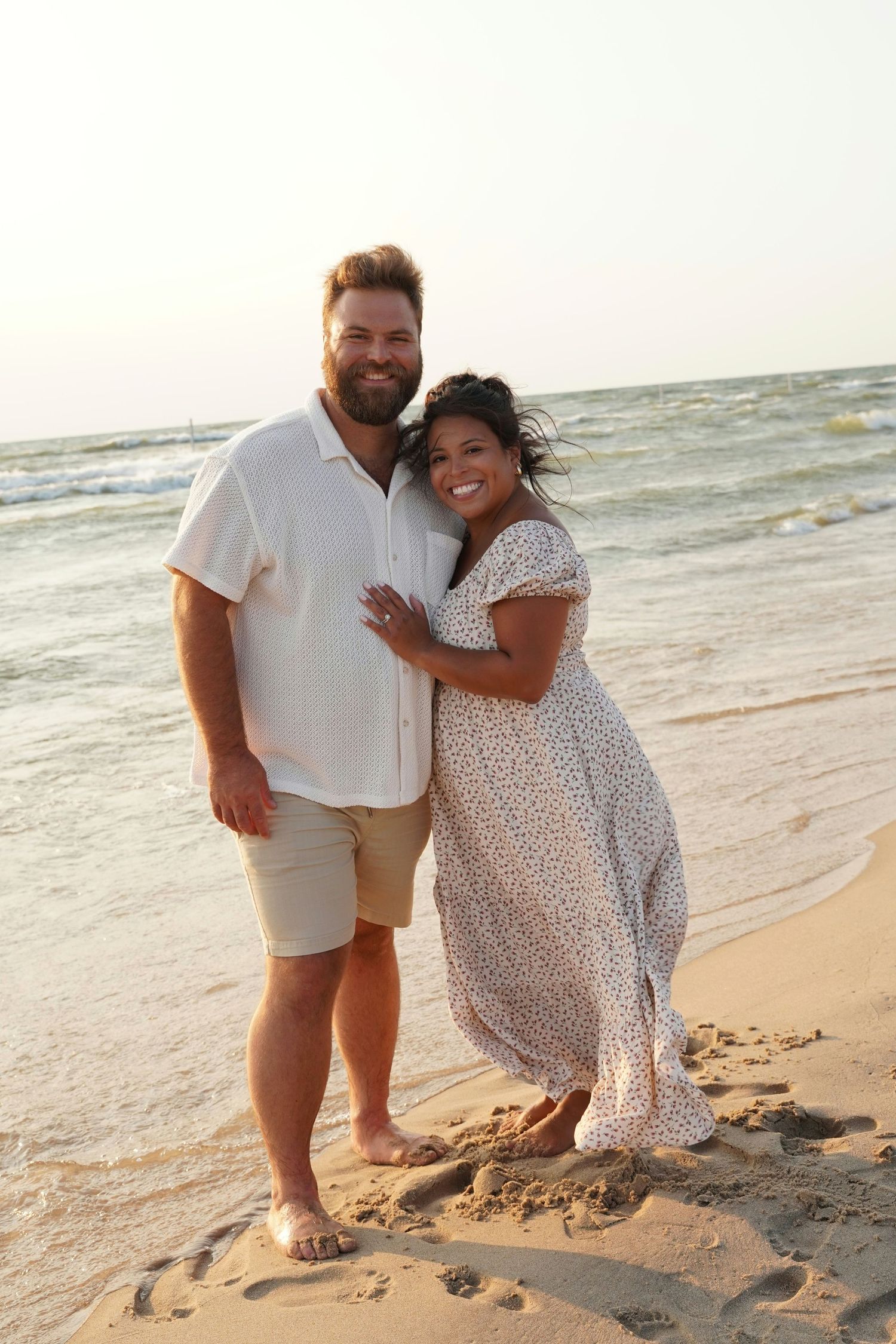 A couple embraces and smiles on a sandy beach at sunset, woman in floral dress, man in white shirt and khaki shorts.
